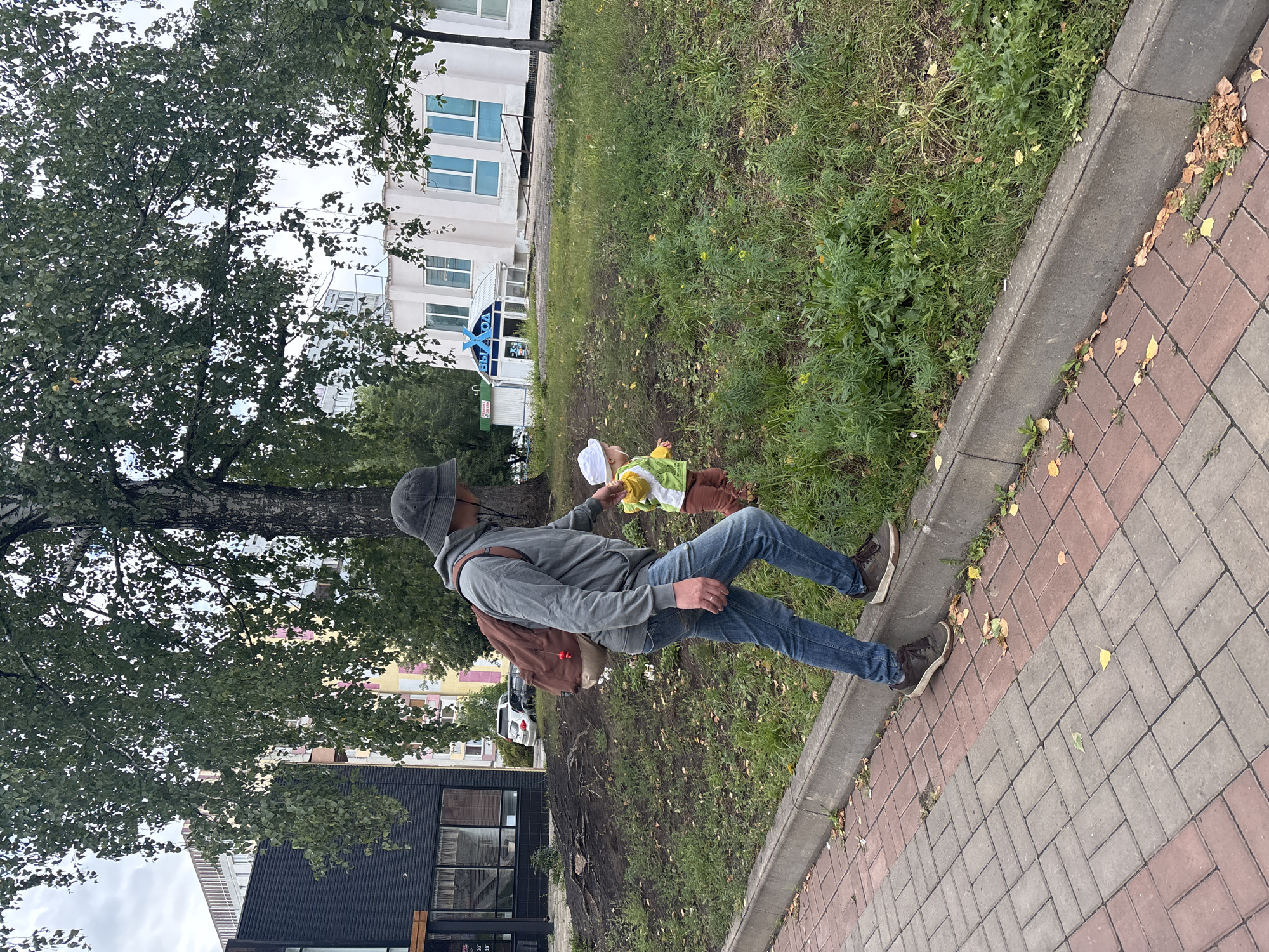 A man wearing a grey hoodie, blue jeans, a dark bucket hat, and a brown backpack is walking on a paved sidewalk next to a curb. He is reaching out to a small child, who is wearing a white hat, a green and yellow shirt, and brown pants, and is standing on the grass next to the curb. Behind them are trees, buildings, and a cloudy sky. The scene appears to be an urban environment in Naberezhnye Chelny, Russia, given the architecture and potential signage in the background, although specific identifiable landmarks are not clearly visible. The overall atmosphere suggests a casual outdoor stroll.