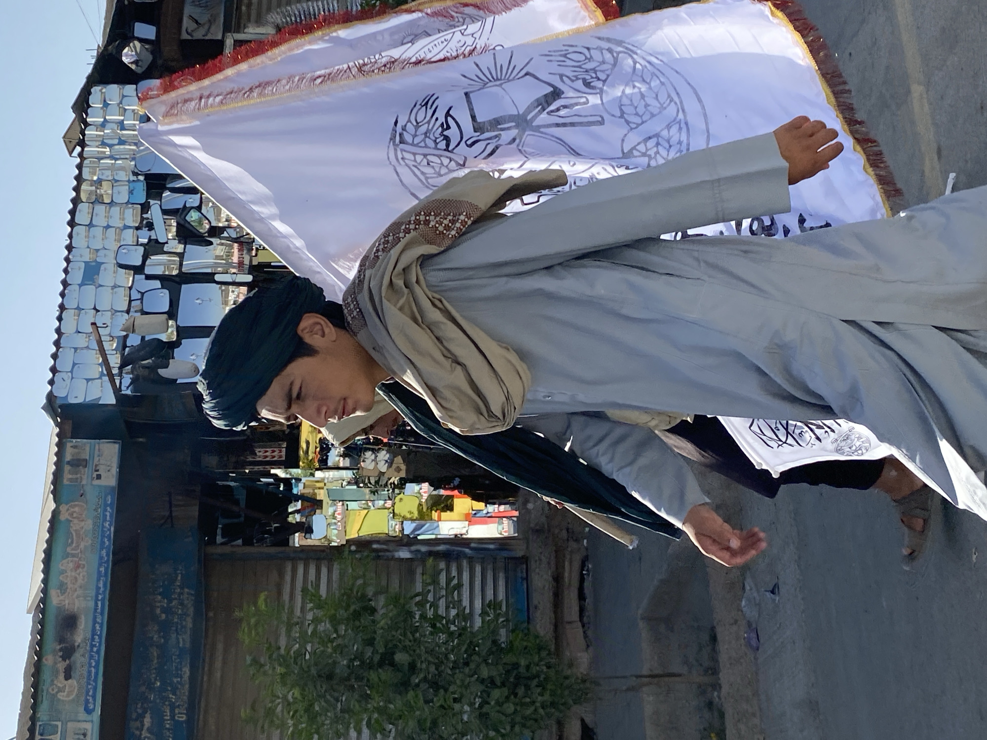 In the city of Kandahar, Afghanistan, a young man wearing a dark green turban and a light blue kameez walks beside a large white flag emblazoned with black Arabic calligraphy. He is also draped in a beige scarf. Behind him, another person in a dark garment is partially visible. The flag has a decorative red and gold fringe along its edge. They are walking on a paved street. To their left, there is a building with a blue awning and shuttered windows. Above the awning, a sign with blue text and images is visible. The building's facade is decorated with numerous reflective square mirrors. A small green tree stands to the left of the building. The sky is clear and blue.