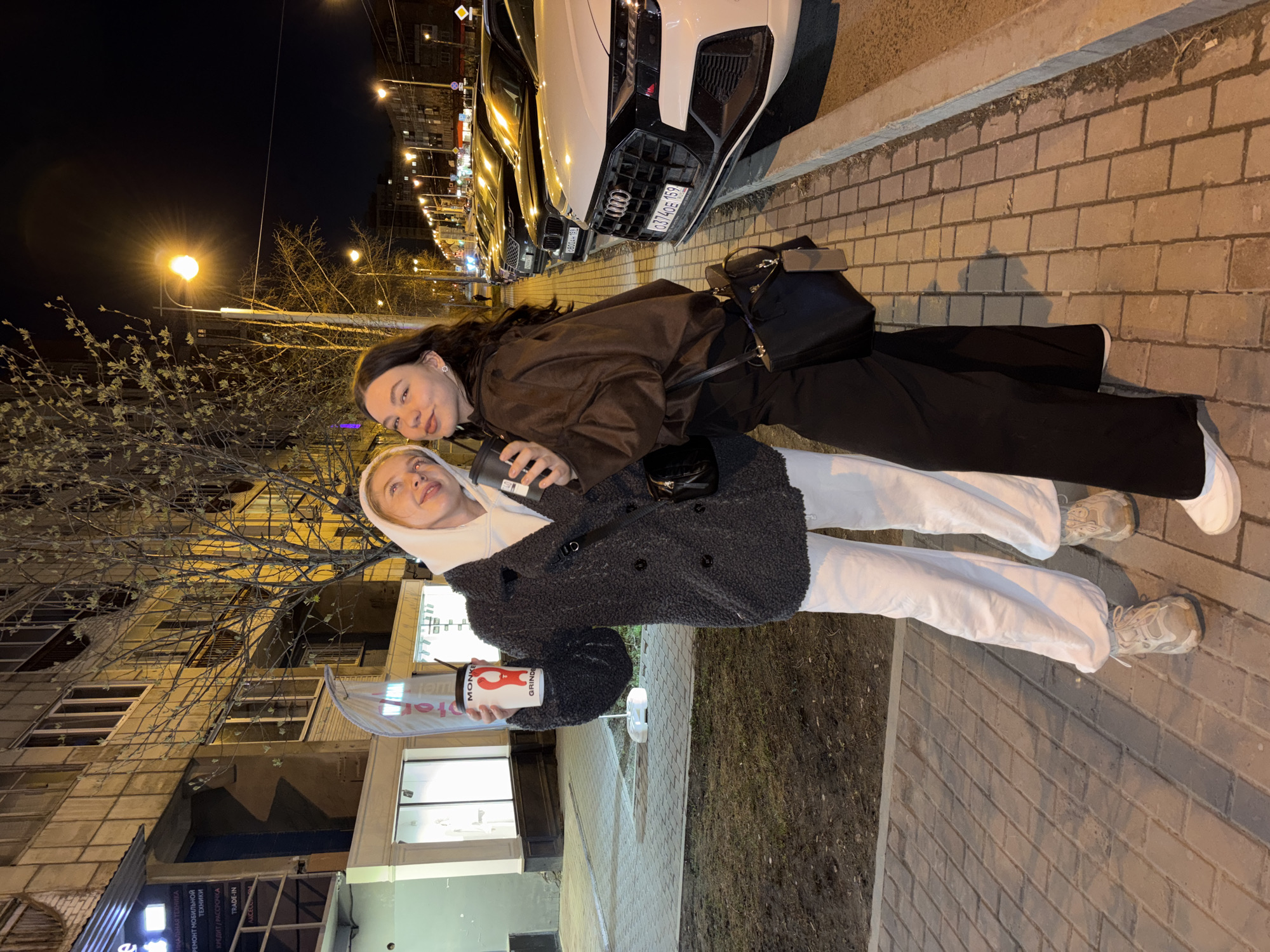 This image captures two young women posing on a city street at night in Perm, Russia. The scene is illuminated by streetlights, indicating evening or nighttime, and the ground appears dry. Bare trees with budding leaves suggest it might be late winter or early spring.
The woman on the right has dark hair and is wearing a dark brown jacket, dark pants, and white sneakers. She carries a black shoulder bag and holds a dark-colored cup. The woman on the left, with blonde hair, is wearing a light-colored hoodie, a textured dark gray coat, white pants, and light-colored sneakers. She holds a white and red cup. Both women are smiling and looking towards the camera, suggesting a friendly interaction or photo opportunity.
In the background, several cars are parked along the street, including a white Audi with a Russian license plate "0374OE 159" (Perm Krai region). Behind the women are multi-story buildings, some with visible storefronts.
Visible text in the image includes:
- On storefront signs: "КАЛЬНАЯ ТЕХНИКА", "РЕМОНТ МОБИЛЬНОЙ ТЕХНИКИ", "КРЕДИТ / РАССРОЧКА", "TRADE-IN", and "AKCELC".
- On the white and red cup: "MONKE GRINDE".
- Partially visible text on the dark cup appears to be "LD".