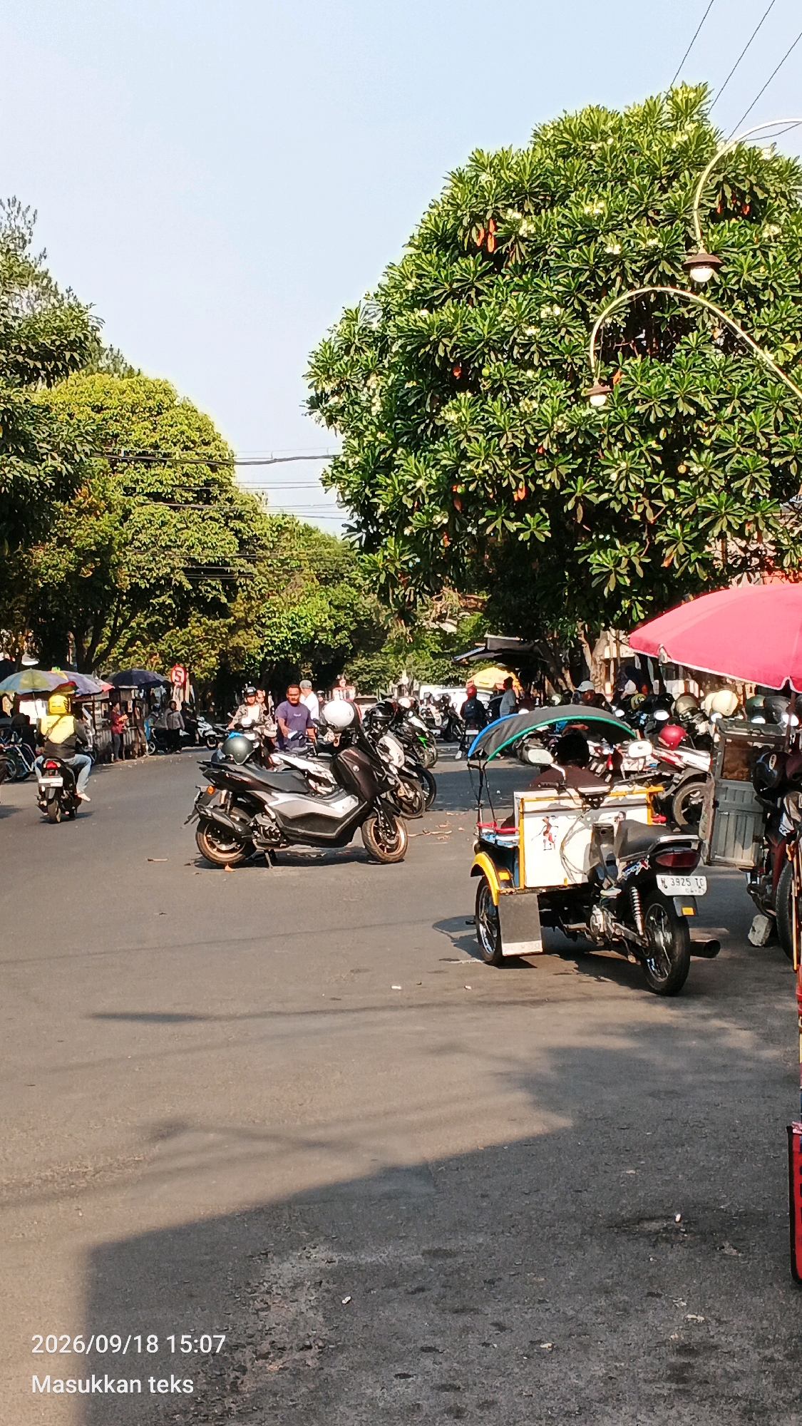 A street scene in Malang, Indonesia, on September 18, 2026, at 15:07. The image depicts a busy intersection with several motorcycles and a three-wheeled vehicle. Lush green trees with visible fruit or seed pods overhang the road, and a clear sky is visible above. Streetlights are mounted on ornate poles. People are visible on and around the vehicles, some appearing to be vendors or shoppers. The road surface shows signs of wear and tear, with shadows cast by the trees and vehicles indicating a sunny day. The overall impression is of a lively, sun-drenched urban environment.