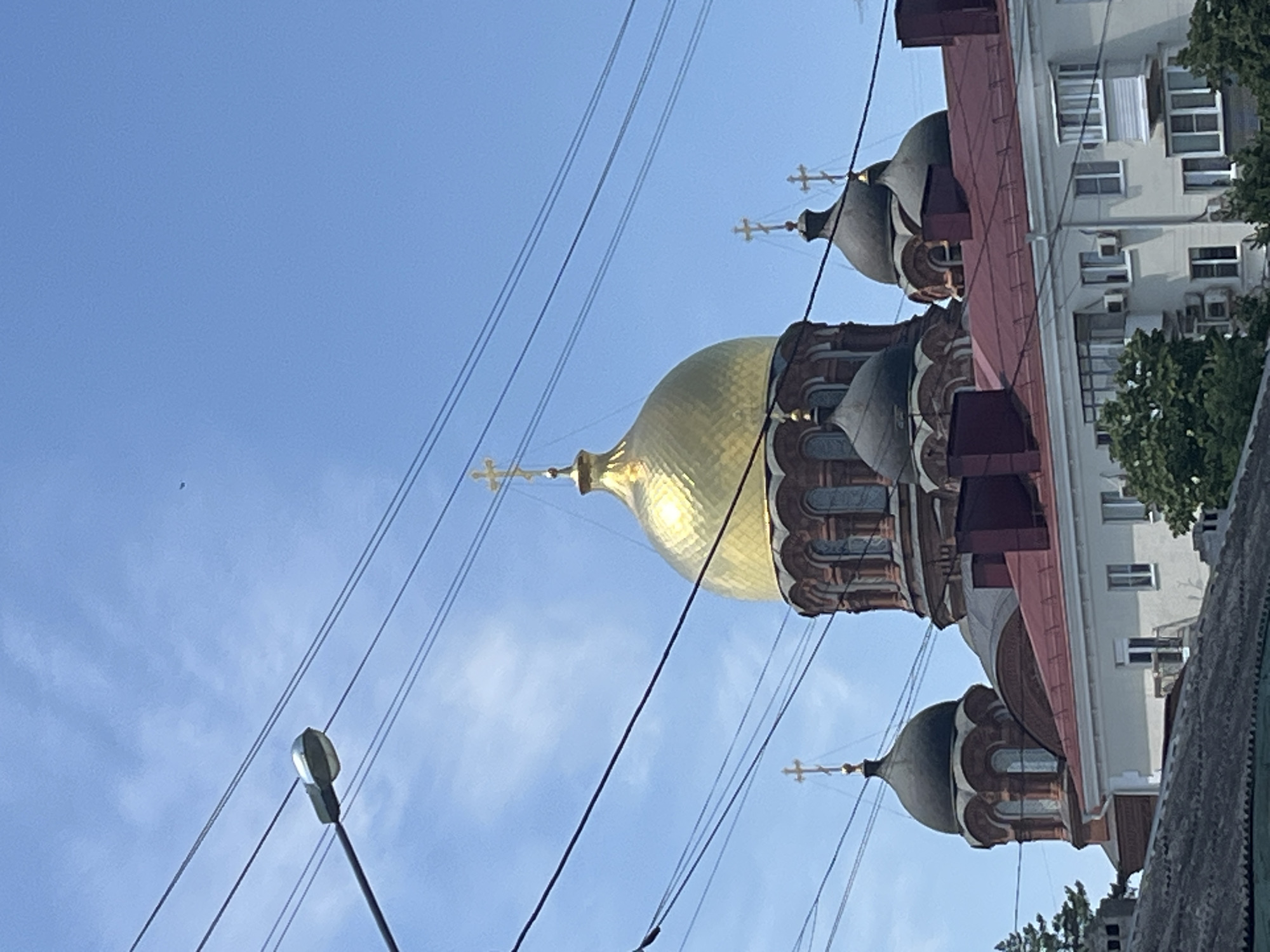 In the city of Krasnodar, Russia, a clear blue sky with sparse clouds is visible. Multiple power lines crisscross the frame, obscuring parts of the scene. In the mid-ground, the golden dome of a large, red-brick Orthodox church stands prominently, topped with a cross. Several smaller, dark-domed cupolas, also topped with crosses, surround the main dome. In the foreground, the roof of a modern apartment building with a red tiled roof is visible, featuring windows and balconies. The overall impression is an urban landscape with significant religious architecture dominating the skyline.
