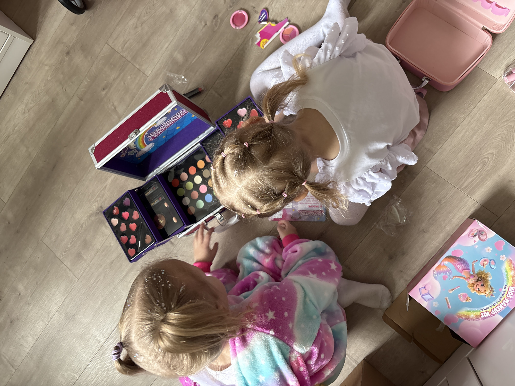 Two young girls are on a wooden floor indoors, engaged with a children's makeup kit. One girl, wearing a white top with a ruffled collar and white tights, is positioned at the top of the frame. The other girl, located at the bottom, is wearing a pink and blue tie-dye pajama set. A large, open makeup case sits to the left, revealing various cosmetic colors. A smaller, pink makeup case is located above the girl in white. A box with "KIDS MAKEUP KIT" written on it sits to the lower right. The overall setting seems to be a child's play area. Given the provided metadata context, the location is Krasnoyarsk, Russia.