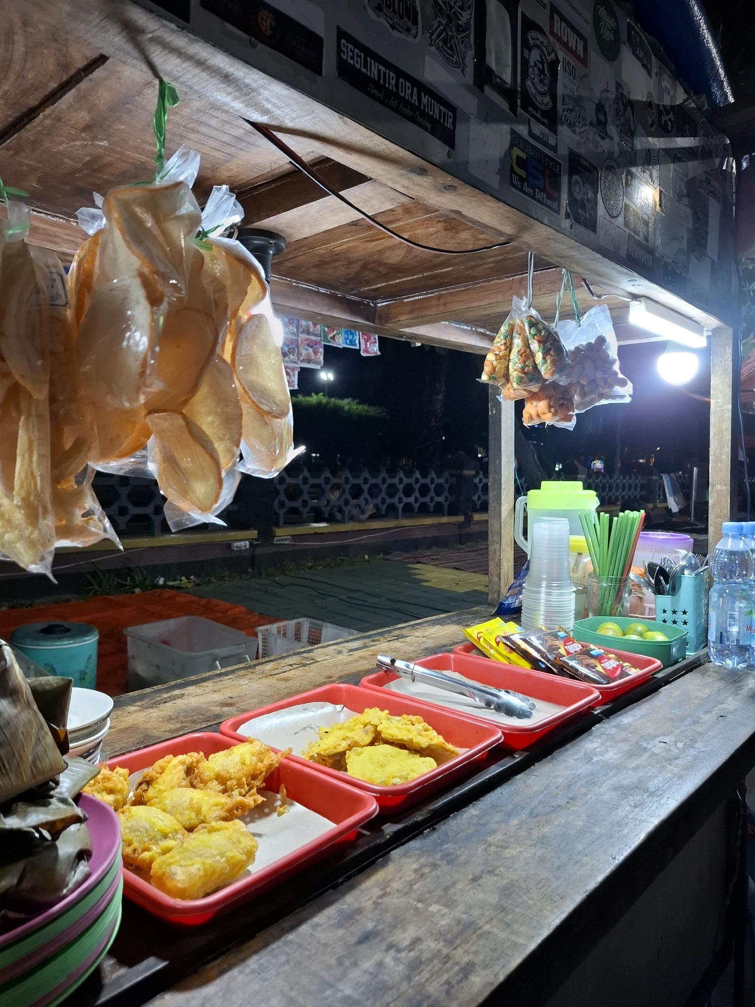 The image depicts an outdoor food stall at night in Pandean, Indonesia. The stall is made of wood and is illuminated by a fluorescent light. Several plastic bags filled with snacks and fried food items are hanging from the ceiling. In the foreground, red plastic trays hold various fried foods, including what appear to be fried bananas and fritters. To the right, there are containers of limes, a pitcher of water, plastic cups, and straws, along with bottles of water and packaged snacks. A blue bin and stacked bowls are visible on the left side. The background shows a dark outdoor area with some fencing and vegetation. The overall scene suggests a bustling night market or street food vendor.
