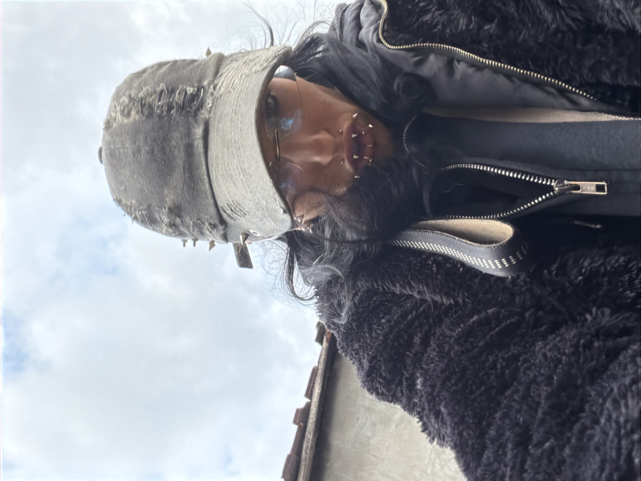 A low-angle, close-up outdoor portrait captures an individual under a cloudy sky. The subject, looking downward towards the camera, has long, dark hair visible beneath a distressed, dark grey baseball cap adorned with metal spikes and grommets. Rimless eyeglasses with tinted lenses are worn. The individual exhibits extensive facial piercings, including a septum ring and multiple vertical labret piercings featuring silver-colored, spike-shaped jewelry on both upper and lower lips, creating a prominent "fang" appearance. A small hoop earring is visible on the left earlobe. The subject is dressed in layered garments, comprising an outer black shaggy faux fur jacket over a black puffer jacket, which is partially unzipped, revealing another dark garment underneath. A dark, textured strap, possibly from a bag, is visible over the left shoulder. The background features a grey concrete or stucco wall in the lower left, topped by a partial terracotta-tiled roofline, set against a predominantly white and grey cloudy sky. While these architectural elements are consistent with structures found in the city of Sofia, Bulgaria, the specific location within Sofia cannot be definitively identified from the visual evidence. The subject is static, posing for the photograph.