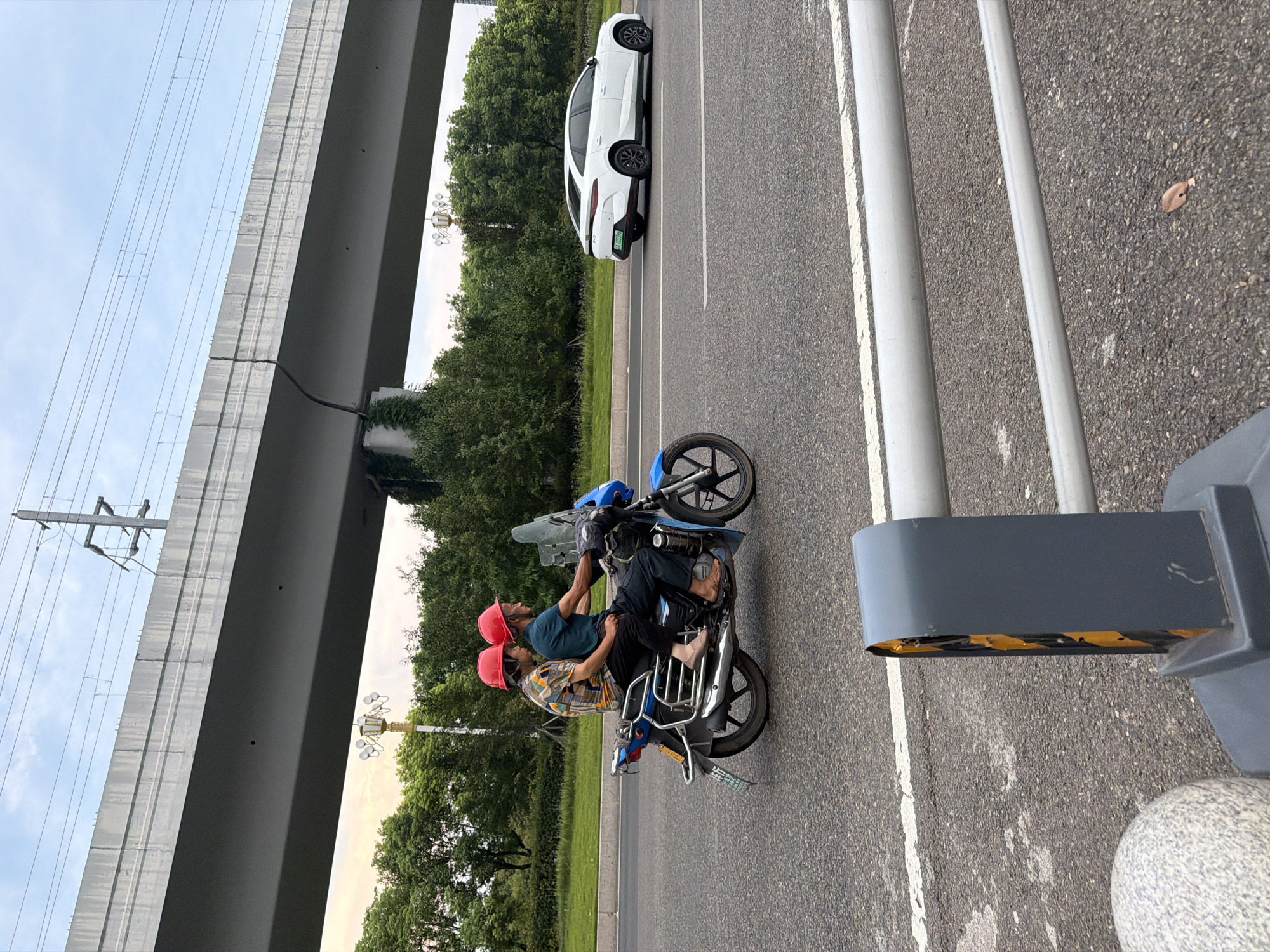 Two individuals, wearing identical red helmets, are riding on a blue motorcycle. The driver, in a patterned shirt, operates the vehicle, while the passenger, in a dark shirt, sits behind them. They are traveling on an asphalt road, indicated by white lane markings. In the background, a modern white car drives in the same direction. An elevated concrete structure, possibly a bridge or overpass, spans the upper portion of the image, with overhead power lines visible against a cloudy sky. A dense line of green trees borders the road, suggesting a suburban or semi-urban environment. A lamppost with multiple lights stands near the trees. A traffic bollard with reflective yellow and black markings is positioned in the foreground on the right side of the road. The scene appears to be set in Jiangjin, China, given the context provided.