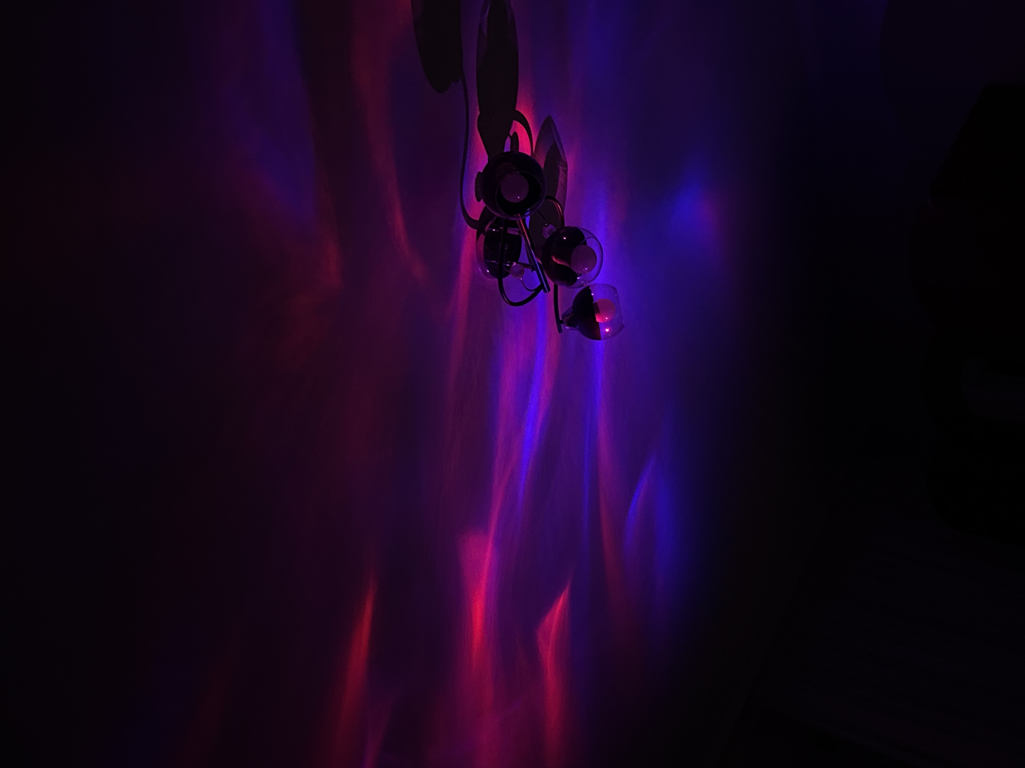 A ceiling fan with three globes, illuminated by pink and purple light, is suspended from a dark ceiling. The light projects ethereal, wave-like patterns across the ceiling and walls, creating an abstract, moody atmosphere. The scene is devoid of human presence, with the focus solely on the interplay of light and shadow. There are no identifying markers of any specific location within the city of Tiraspol, Moldova.