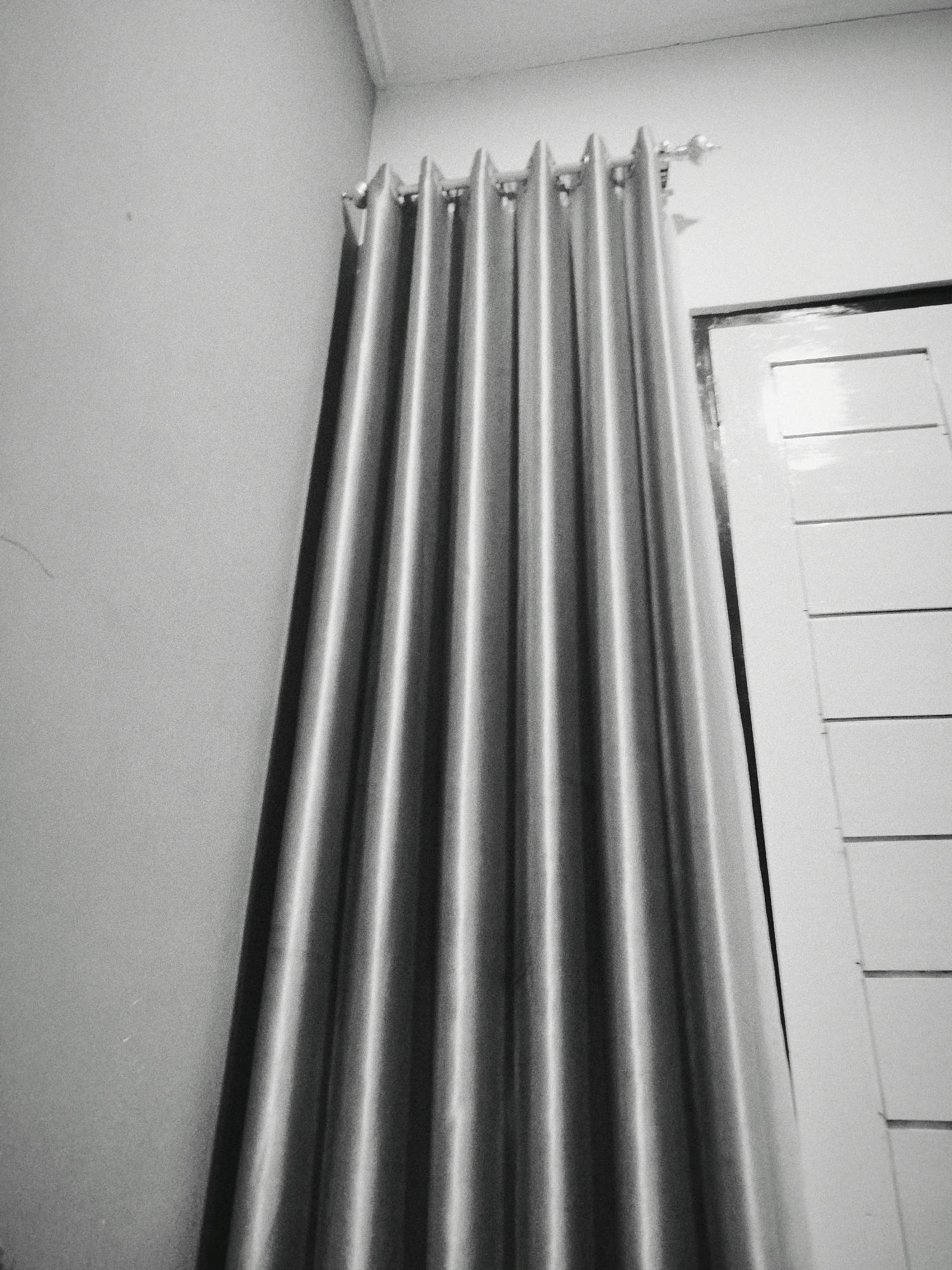 A black and white, medium shot, indoor photograph shows a partially drawn, silver curtain hanging from a rod. The curtain is pleated and falls vertically, obscuring a portion of a white paneled door to the right of the frame. The wall to the left of the curtain is a light gray, with a visible seam near the top of the frame where it meets the ceiling. The overall impression is of a domestic interior, potentially a bedroom or living area. No people or animals are present.