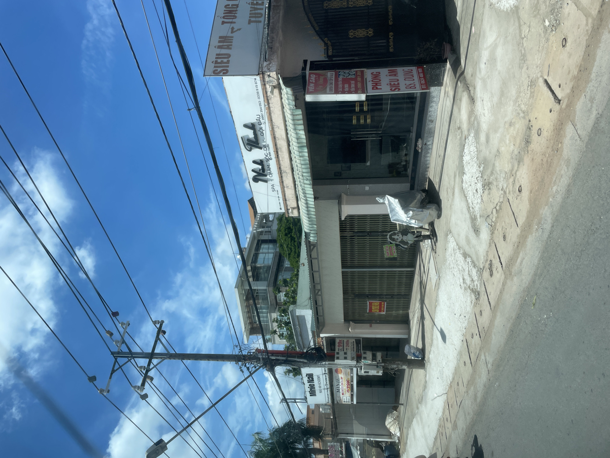 The image shows a street scene in Phường An Tịnh, Vietnam, under a bright blue sky with scattered clouds. Utility poles with numerous wires are prominent in the foreground and midground, crisscrossing the sky. Along the street, there are several businesses. To the left, a building with a sign reading "Minie Nail" is visible, with smaller signs below advertising prices. Further to the right, a building with a large glass storefront has signage that includes "Nha Thanh SPA" and "Chăm Sóc Da" (Skin Care). Adjacent to this is another building with a sign in Vietnamese that appears to be for a "Phòng Siêu Âm" (Ultrasound Room). A gate made of gold-colored metal is to the far right. The street in front of the businesses is paved with concrete and asphalt, with some areas showing wear and tear. A covered object resembling a motorcycle or scooter is parked on the sidewalk. Various trash receptacles and smaller signs are also present. A modern multi-story building with balconies and glass windows is visible behind the street-level businesses.