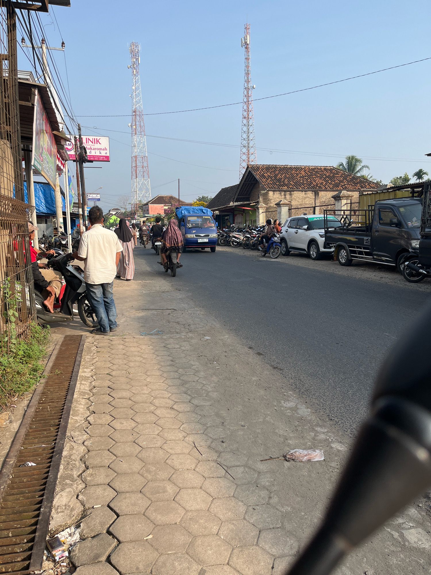 The image depicts a street scene in Kemuning, Indonesia, on a clear day. In the foreground, a grey pickup truck is parked on the right side of the road. To its left, a white SUV is also parked. The road is paved, and a sidewalk made of hexagonal pavers runs alongside it. A metal grate drain is visible along the edge of the sidewalk.
Several motorcycles and their riders are present on the road and sidewalk. One person, wearing a white shirt and blue jeans, stands on the sidewalk. Another person in a long dress and headscarf stands nearby. Further down the road, other individuals are seen on motorcycles.
In the background, buildings with tiled roofs are visible. Two prominent cell towers stand tall against the blue sky. Signs are posted on the buildings, including one that reads "KLINIK NUR RAHMAH". The overall impression is of a busy street in a town or city.