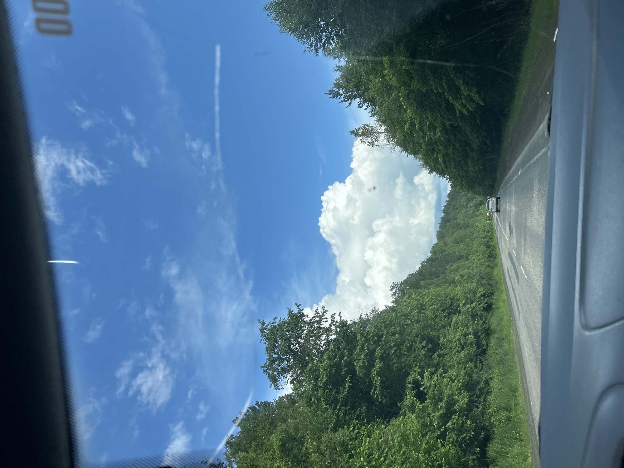 This is a photo of a road in the city of Usman-Gali, Russia, on a cloudy day. The road is paved and has two lanes of traffic. There are trees on either side of the road. The sky is blue with some white clouds. The photo appears to have been taken from inside a car. There are other cars on the road, but no people are visible.