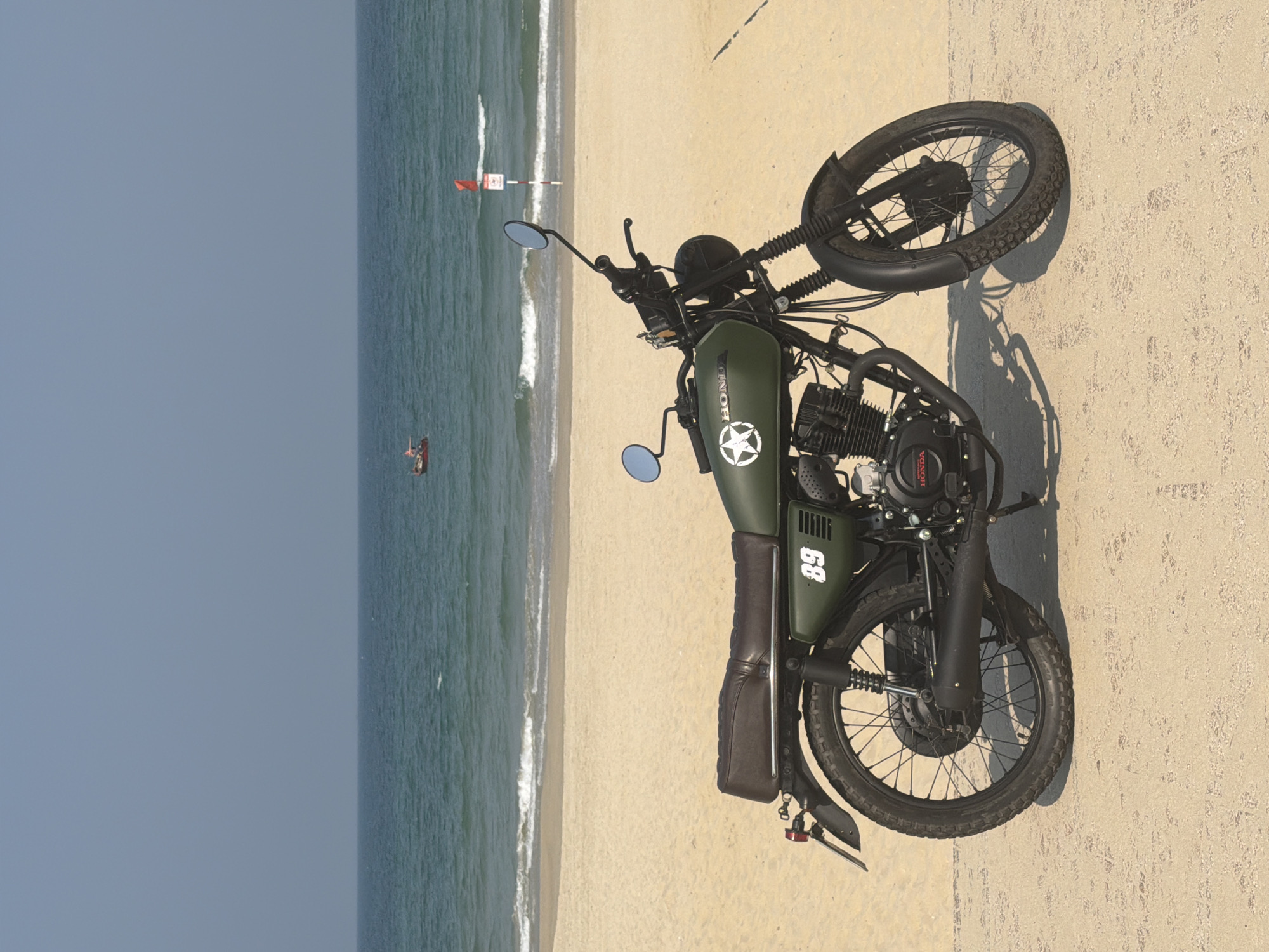 A horizontal, medium shot shows a green, vintage-style Honda motorcycle with a white star emblem, parked on a tan, sandy beach. The number "89" is displayed on the motorcycle. The motorcycle is the primary subject, its frame and features clearly visible.
Behind the motorcycle is a blue ocean, with gentle waves lapping onto the shore. In the distance, a red flag on a pole marks a swimming safety zone. A small boat with a person is further out in the ocean. The sky above the ocean is a light blue, suggesting a clear day.
The scene depicts a serene coastal environment, with the motorcycle seemingly enjoying a moment of rest on the beach. Based on the location and vegetation, it may be one of the beaches of Đà Nẵng, Vietnam, known for its long sandy shorelines and clear waters. There is no direct evidence to confirm this is Đà Nẵng but the beach and the vegetation are consistant with this location.