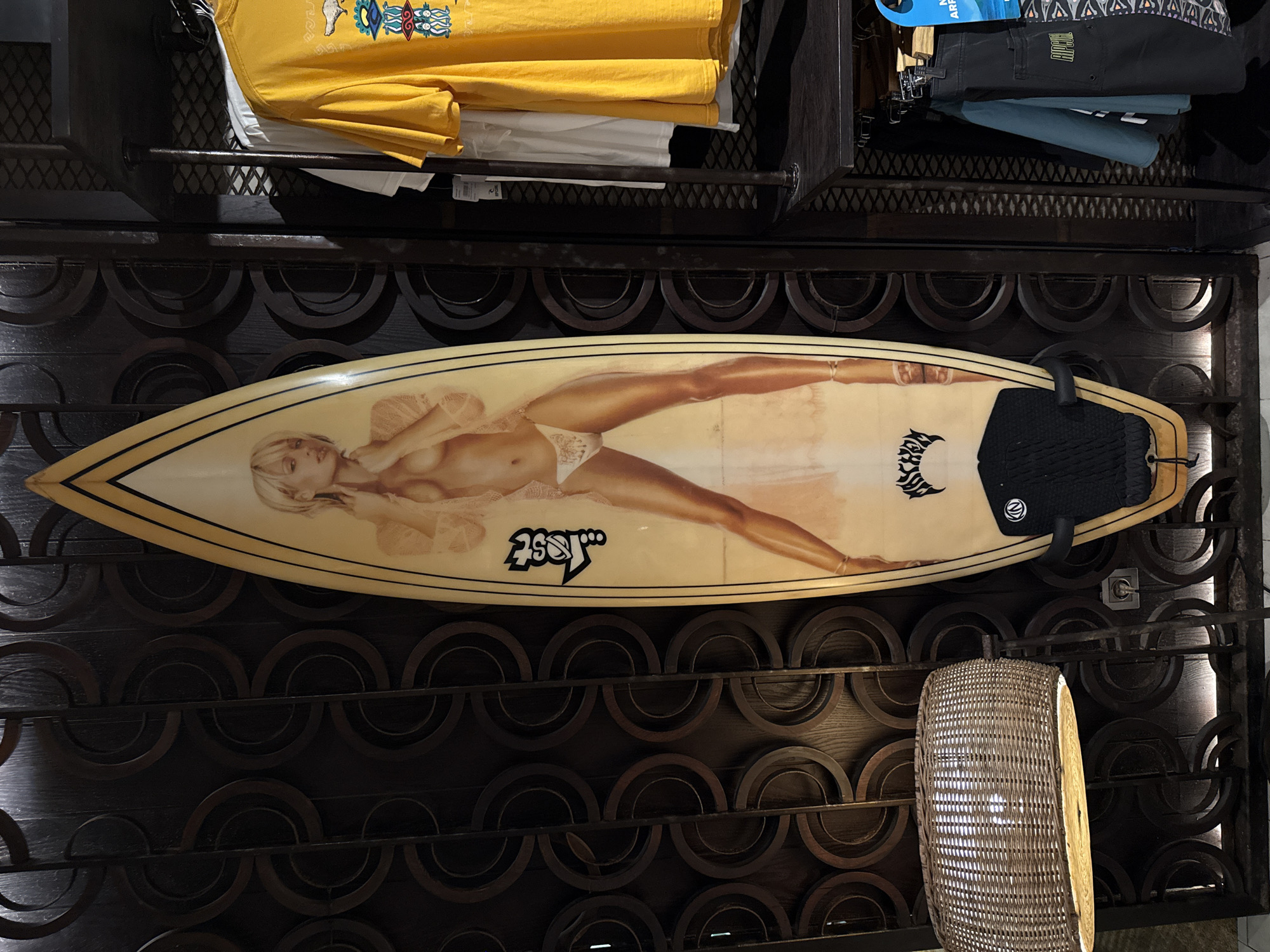 The image depicts a surfboard displayed on a dark, decorative shelving unit in what appears to be a retail store. The surfboard has a light tan base color with an image of a blonde woman in minimal clothing airbrushed onto its surface. There are also logos and branding visible on the surfboard. Above the surfboard, there are clothes hanging on a metal rack, including a yellow t-shirt. Below the surfboard, there is a woven basket-like object. The background appears to be a store setting, which might be located in Kuta, Indonesia, considering the surfboard's likely association with surfing culture and the presence of retail clothing. There are no people visible in the frame.