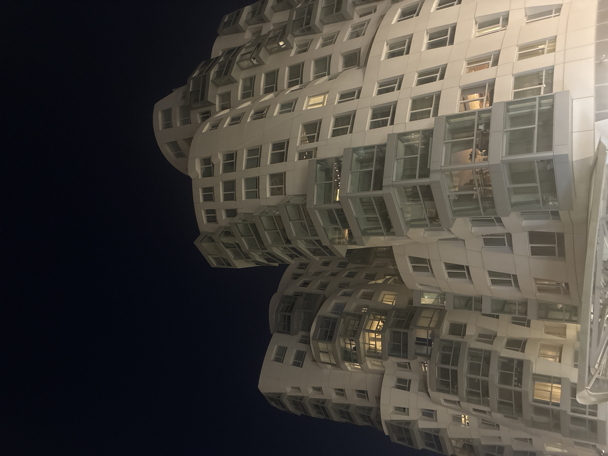 Here is a description of the image:
A low-angle, nighttime photograph shows a contemporary apartment building located at 100 Bishopsgate, London, United Kingdom, known for its distinctive stacked box-like design. The building has many windows and external glass cubicles, some of which are illuminated from within. The building is primarily white or light gray. A dark blue sky occupies the left of the image.