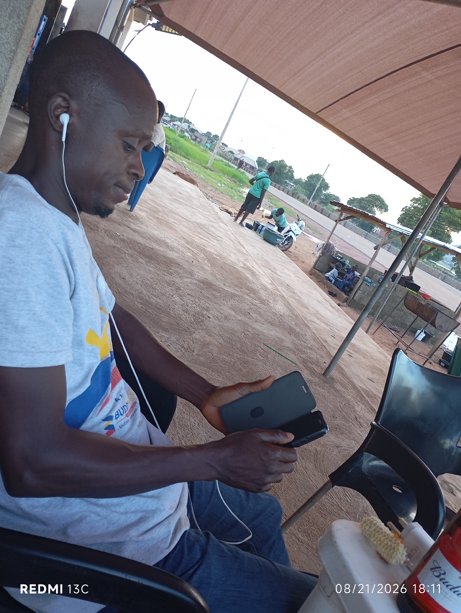 A man with short hair and an earpiece is sitting in a chair, holding two cell phones. He is wearing a grey t-shirt with a colorful logo and blue jeans. In the background, several people are gathered near a motorcycle and small structures with corrugated metal roofs, suggesting an outdoor market or roadside stop. The ground is dusty and uneven. The scene is likely outdoors, possibly in a town or village setting in Shendam, Nigeria, given the architecture and general environment. A bottle of Budweiser and a partially eaten corn cob are visible on a white surface in the foreground. The timestamp "08/21/2026 18:11" and "REDMI 13C" are superimposed on the image.