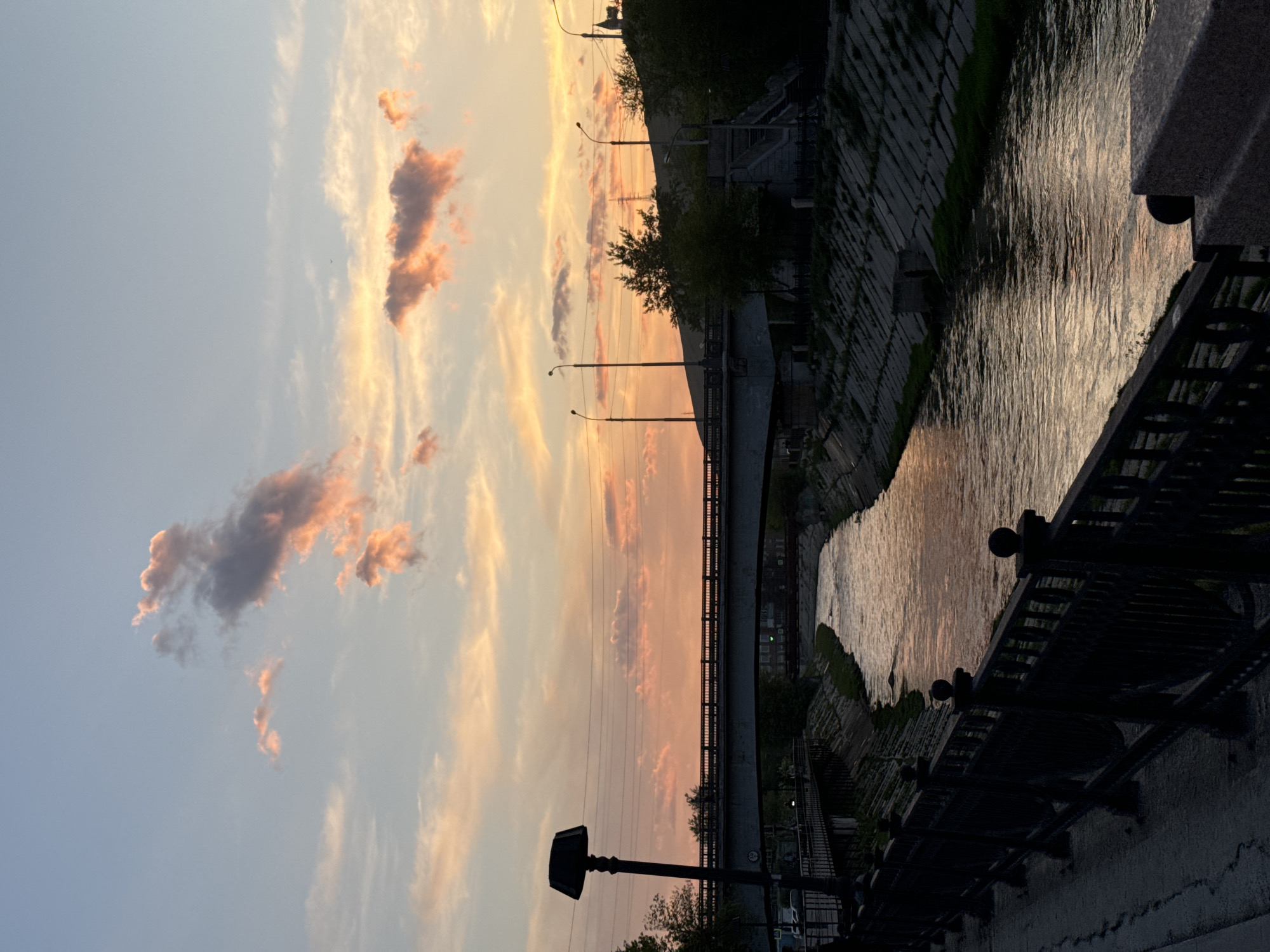 The image is an eye-level, outdoor shot capturing a sunset over the Yenisey River in Krasnoyarsk, Russia.
The sky is a gradient of pale blue to yellow and pink hues, with scattered clouds catching the light. Vertical power lines bisect the sky. Below the sky, a bridge spans the river. Street lamps are visible along the walkway.
On the right side of the frame is the riverbank, constructed of stone. A wrought iron fence runs along the embankment. The water reflects the colors of the sky. The image is slightly underexposed.