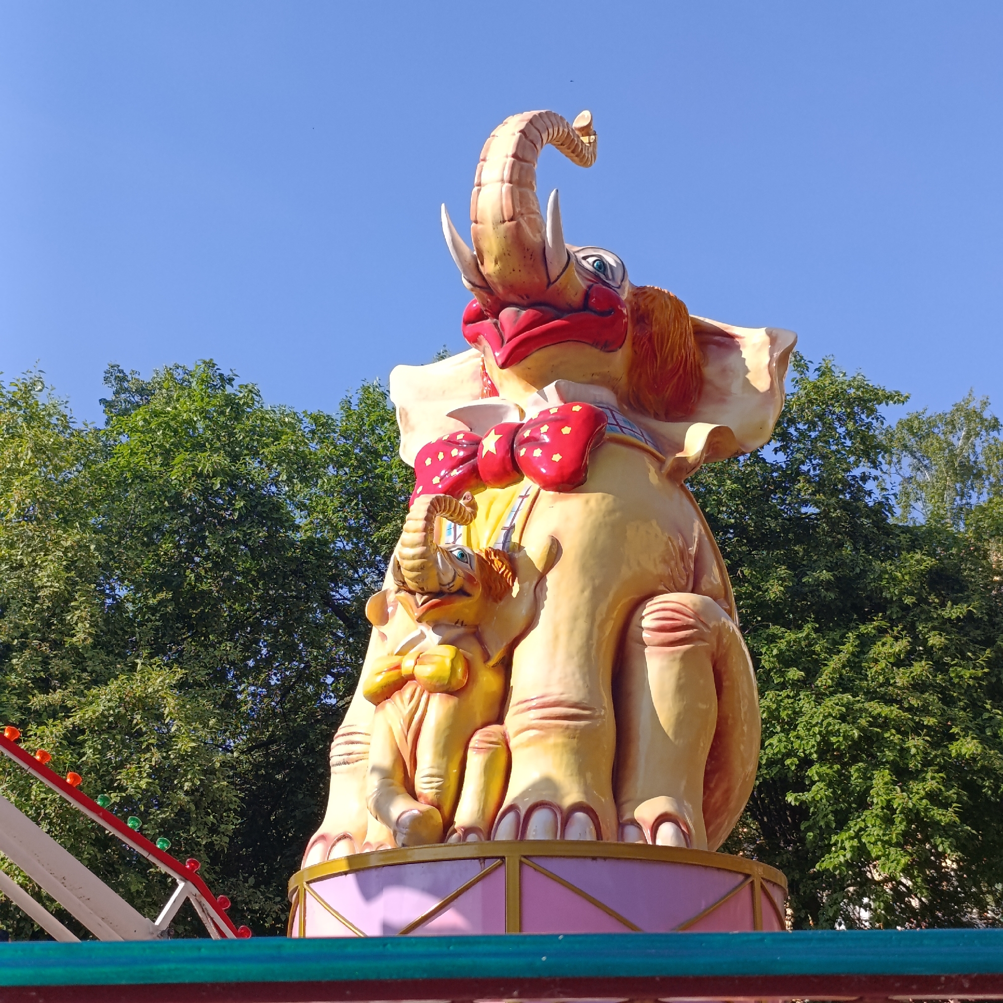 An outdoor scene features a colorful, cartoonish elephant sculpture. The elephant is primarily yellow, with red lips and a red bow tie with yellow stars. A smaller elephant with a yellow bow tie clings to the larger elephant's body. The sculpture is placed on a pink and white striped platform. Green trees form the backdrop against a clear blue sky. A portion of a white and red structure with colored bulbs is visible on the left, suggesting this may be part of an amusement park ride. The sculpture is located in the city of Izhevsk, Russia.
