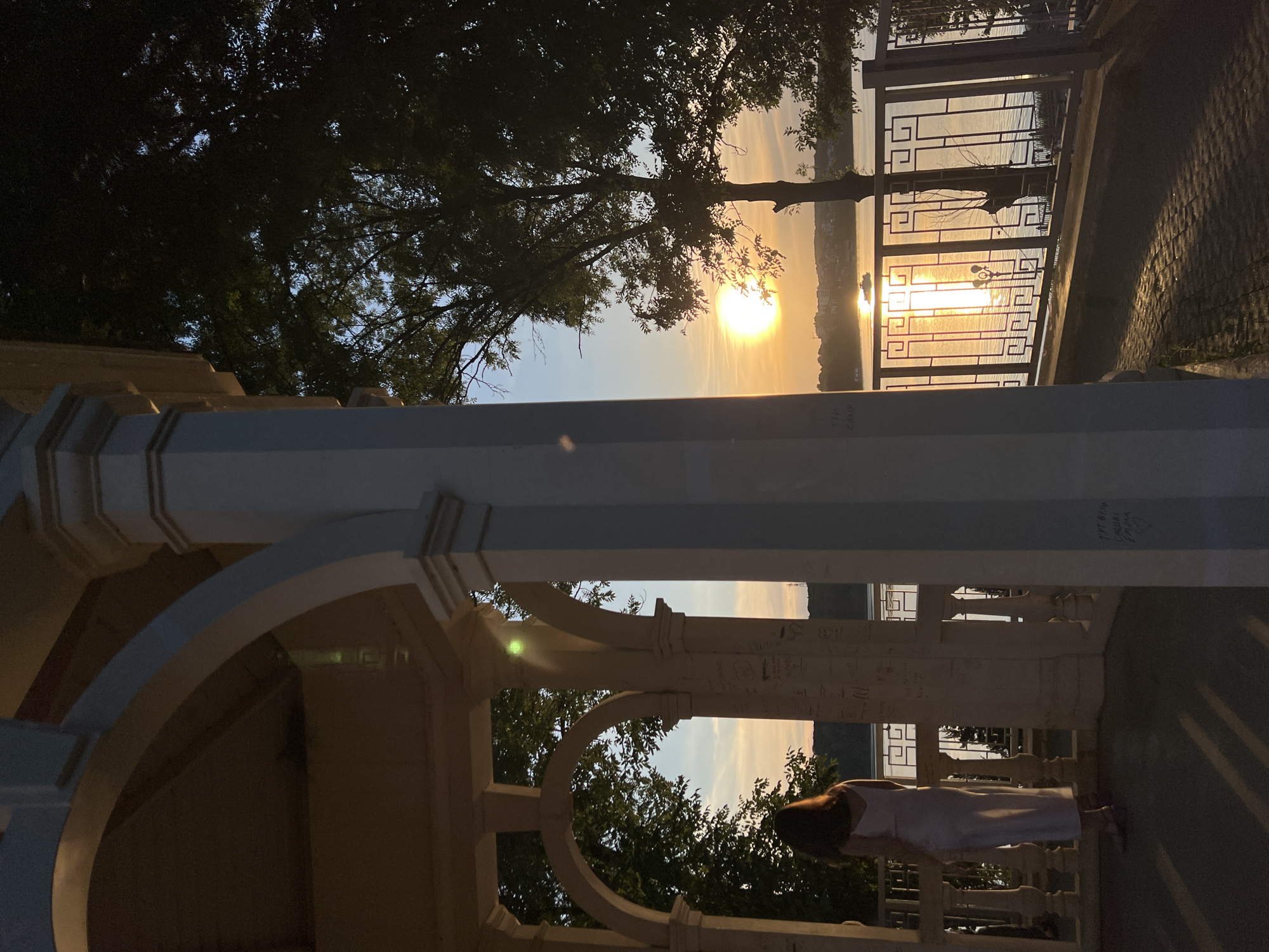The image depicts a sunset over a body of water, likely the Ternopil Pond, in Ternopil, Ukraine. A woman stands on the covered veranda of a white gazebo, her back to the camera, wearing a long white dress. The gazebo features arched openings and white pillars inscribed with graffiti. Through the arches, trees and the pond are visible. The sun, a bright orange orb, is setting on the horizon, casting a warm glow on the water and the sky. A small boat is visible on the pond. A metal railing with a geometric pattern runs along the edge of the veranda overlooking the pond. The ground in front of the veranda is paved with cobblestones, and the lighting suggests the late evening. The overall atmosphere is serene and tranquil.