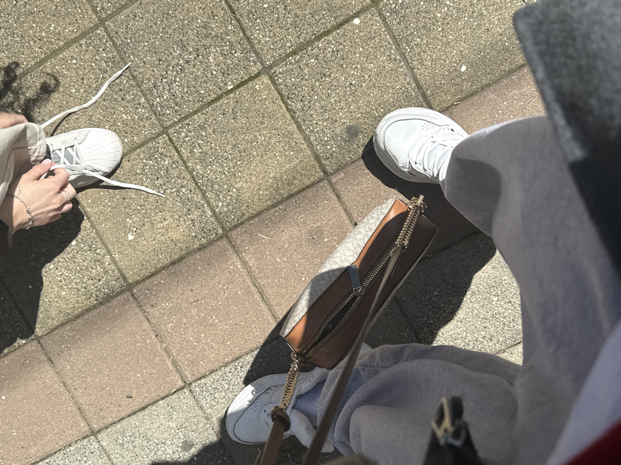 The image presents an overhead view of two individuals standing on a brick-paved surface, likely a sidewalk. On the left, a person is bent over, their hands engaged in tying the laces of a white Adidas Superstar sneaker. A delicate silver bracelet adorns their wrist. The other individual is dressed in grey sweatpants and white sneakers. A brown and beige handbag with a gold chain strap sits near their feet. The location is unidentified.
