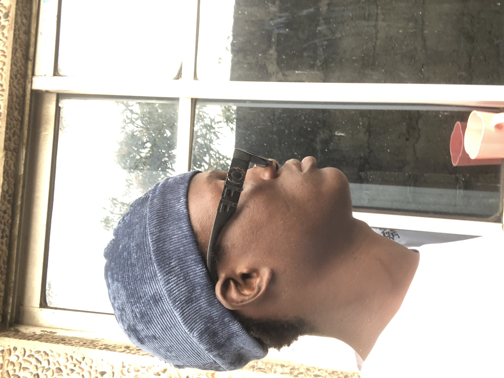 In Ado, Nigeria, a young Black person, viewed in profile from the left, wears a blue beanie and dark sunglasses with a rectangular frame. The person is positioned against a window with a textured, stone-like wall to the left and a white frame on the right. Reflected in the window are trees and a cloudy sky. In the foreground, a portion of a white t-shirt is visible, and two pink mugs are partially seen through the glass.