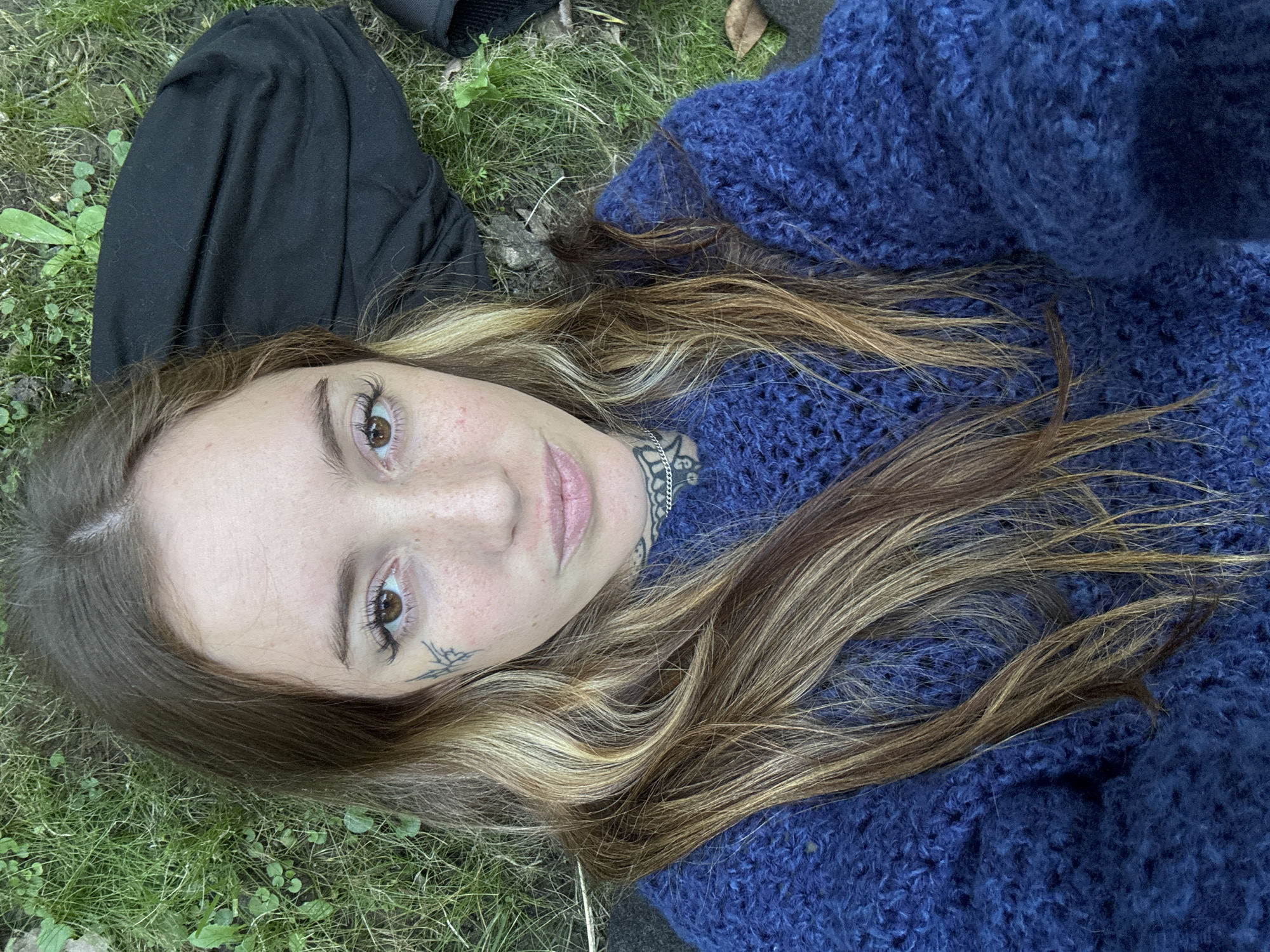 A woman is lying on her back on grass. She is wearing a blue sweater. She has brown and blonde hair, brown eyes, and a tattoo below her left eye and on her neck. Part of what appears to be black clothing is visible to the left. The location is Villa General Belgrano, Argentina.