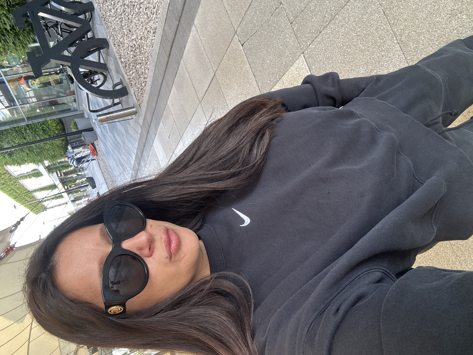 A person with dark hair is wearing black sunglasses and a black Nike sweatshirt. They are sitting or standing on a paved surface in an urban environment. In the background, there is a building with greenery and some metallic structures that may be bicycle racks or public art.