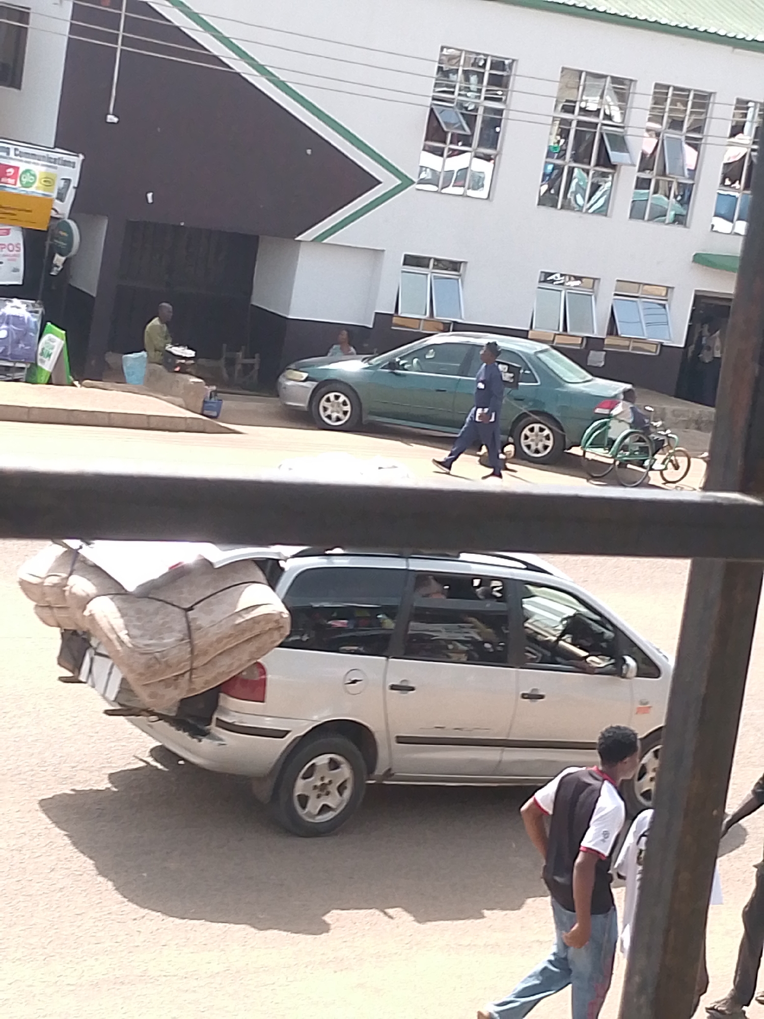 The image depicts a street scene in Bukuru, Nigeria, likely during the day given the shadows. In the foreground, a silver station wagon is heavily loaded with large, bundled items strapped to its rear. The bundles appear to be sacks or similar large packages covered in a light-colored material. A young man in a black and white shirt and blue jeans walks away from the camera, his posture suggesting movement.
In the mid-ground, a dark green sedan is parked on the street. A person dressed in a dark blue uniform walks away from the sedan. Beside the sedan, a person in a wheelchair is being pushed or is operating a manual wheelchair with large spoked wheels. Behind the sedan, the facade of a building is visible. The building is white with dark brown sections and green diagonal accents. It has multiple windows, some of which are open. There are signs on the building, one of which appears to have text on a yellow background and the words "Communications".
In the background, a man sits on a ledge in front of the building, with a blue bucket and some objects on the ledge. The general setting appears to be an urban or semi-urban area with a mix of commercial buildings and street activity. The weather seems clear, allowing for distinct shadows.