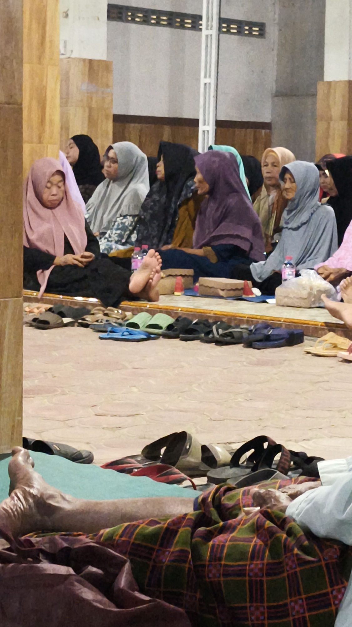 In Dalegan, Indonesia, a group of people, mostly women wearing hijabs of various colors including pink, grey, black, and purple, are seated on a tiled floor, likely inside a mosque or community hall, given the architectural elements visible in the background. Some individuals appear to be listening or praying. In the foreground, a person's legs are visible, covered by a plaid blanket, with a foot resting on a green mat. Several pairs of sandals and flip-flops are neatly arranged on the floor in front of the seated individuals, suggesting a common area where footwear is removed. The lighting is artificial, casting a warm glow on the scene. The overall atmosphere appears to be one of quiet gathering or religious observance.