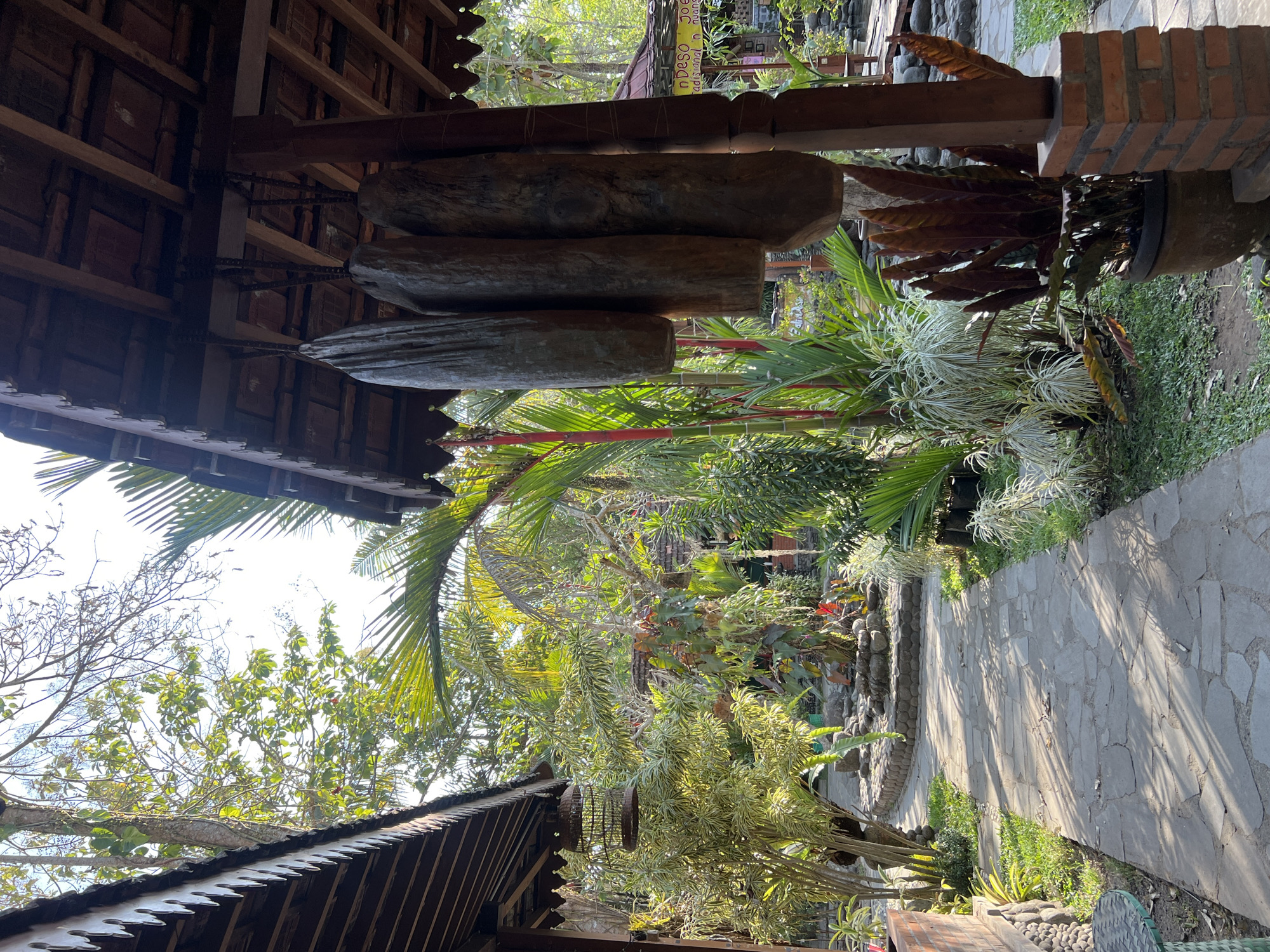 The image depicts a serene outdoor scene in Tlogoadi, Indonesia, likely a garden or courtyard area with traditional architecture. A stone pathway meanders through lush greenery, with various plants and trees flanking its sides. Overhead, a wooden roof with terracotta tiles is visible, supported by wooden posts. Suspended from the roof are three large, weathered wooden objects, possibly decorative or functional items. In the foreground, a brick pillar supports one of the wooden posts. A birdcage hangs from a tree branch to the left, and in the background, hints of other structures and foliage suggest a larger compound. The sunlight casts dappled shadows on the pathway, indicating a bright, possibly morning or afternoon, setting. There are no people visible in the frame.