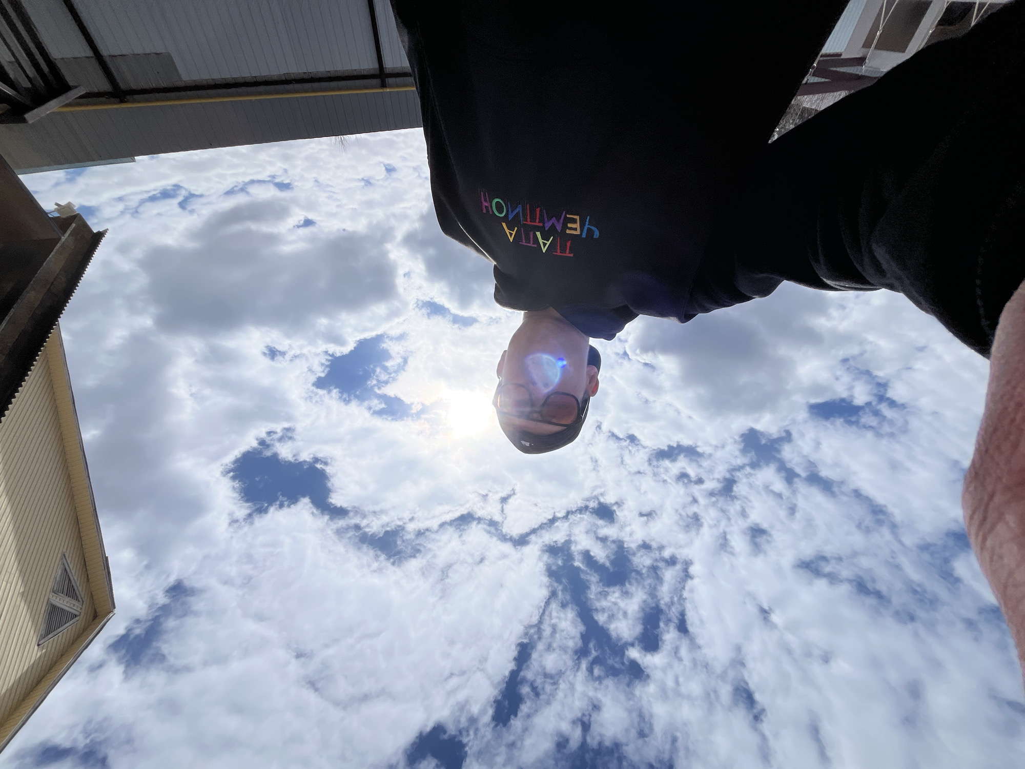 The image is an upside-down selfie of a person wearing a black hoodie with colorful writing. The person is wearing glasses and a black cap. The background features a blue sky with white clouds. A building with light siding is visible on the left, and a metallic structure is visible on the top left. The location is Chaikovsky, Russia, as indicated by the reversed Russian text on the hoodie.