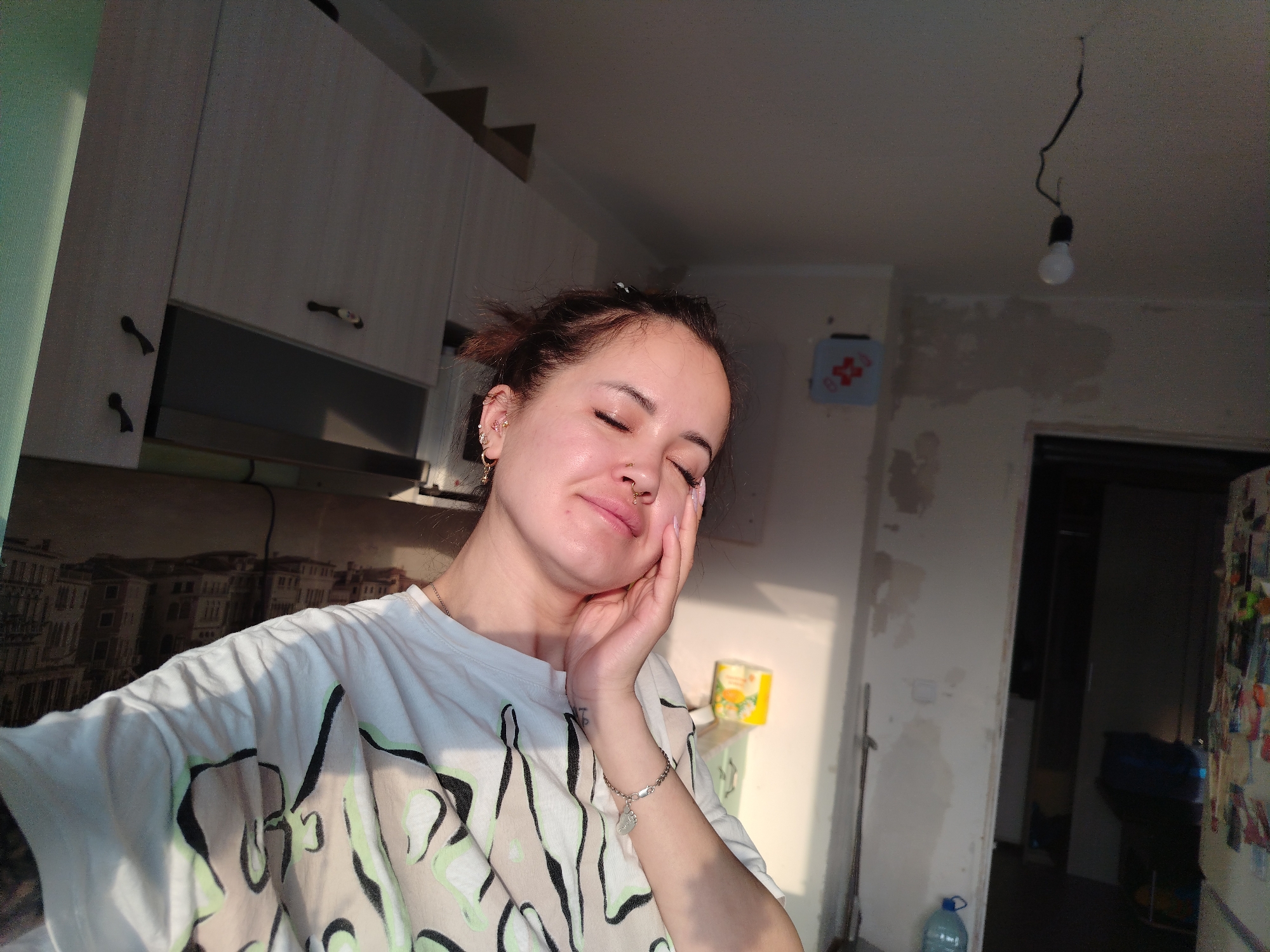 A woman with brown hair pulled back is posing with her eyes closed and her right hand on her cheek. She is wearing a white t-shirt with a graphic design and a silver bracelet. The background includes kitchen cabinets, a partially renovated wall, a bare light bulb hanging from the ceiling, and an open doorway. The image appears to be taken indoors. This image does not contain the location in the city of Noviy, Russia.