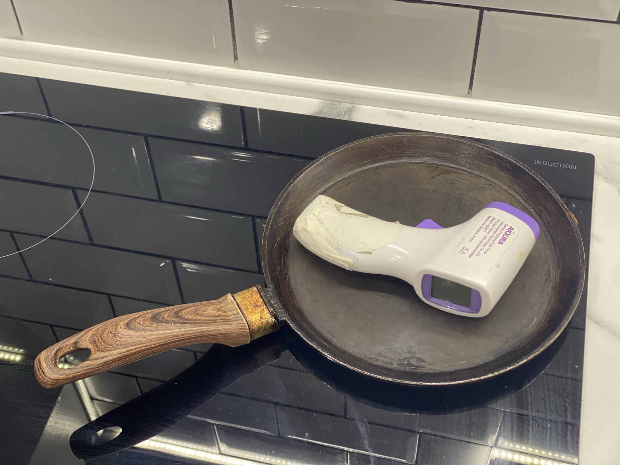 A digital thermometer is placed inside a metal frying pan with a wooden handle. The frying pan is resting on a black stovetop with white tile backsplash. The thermometer is white with a purple trigger and a digital display. The handle of the frying pan has a wood grain pattern. There is some white tape on the thermometer. The scene is indoors, on a kitchen counter. There is no indication of the location being Dolgoprudny, Russia.