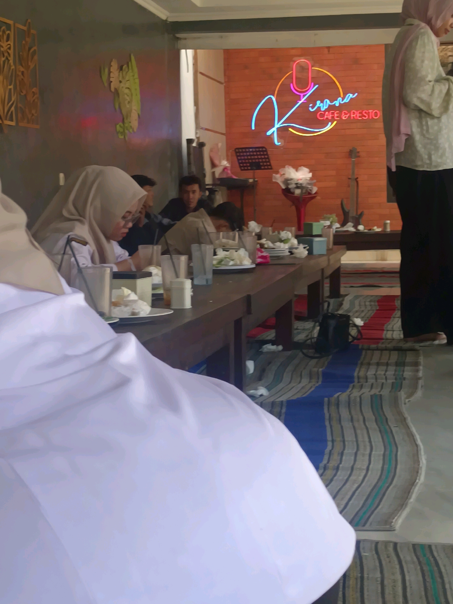The image depicts an indoor scene at the "Kirana Cafe & Resto" in Kerangkulon, Indonesia. The cafe features a brick wall with a neon sign spelling out "Kirana Cafe & Resto" above a microphone graphic. In the foreground, a person wearing white is partially visible, seated at a long wooden table. Other individuals are seated at the table, some wearing hijabs. Various plates of food, drinks in cups, and napkins are on the table. The floor is covered with a striped rug. In the background, a guitar and a red vase with white flowers are visible. The overall atmosphere appears casual and social.