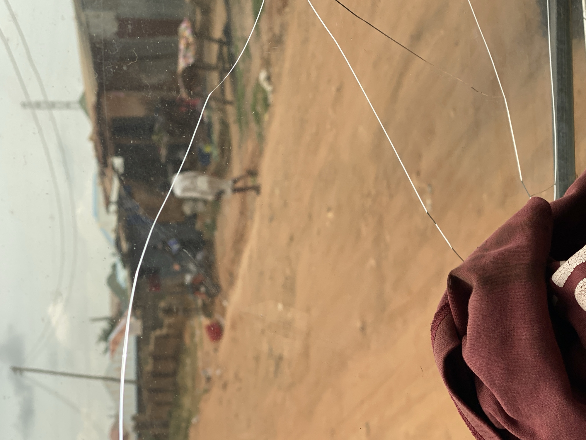 The image depicts a view through a cracked glass pane, likely a window, looking out onto a dusty, outdoor environment. In the foreground, a crumpled piece of burgundy fabric, possibly clothing, is visible. The background shows a dirt road with sparse vegetation and what appear to be simple structures, possibly homes or market stalls, constructed from materials like corrugated metal and wood. A person in light-colored clothing is walking away from the camera in the mid-ground. Electrical wires and a utility pole are present in the distance, indicating an inhabited area. The overall impression is one of a rural or semi-urban setting in Nigeria, given the mention of Barakin Akawo. The cracks in the glass obscure and distort the view of the external scene, adding a layer of fragility to the image.