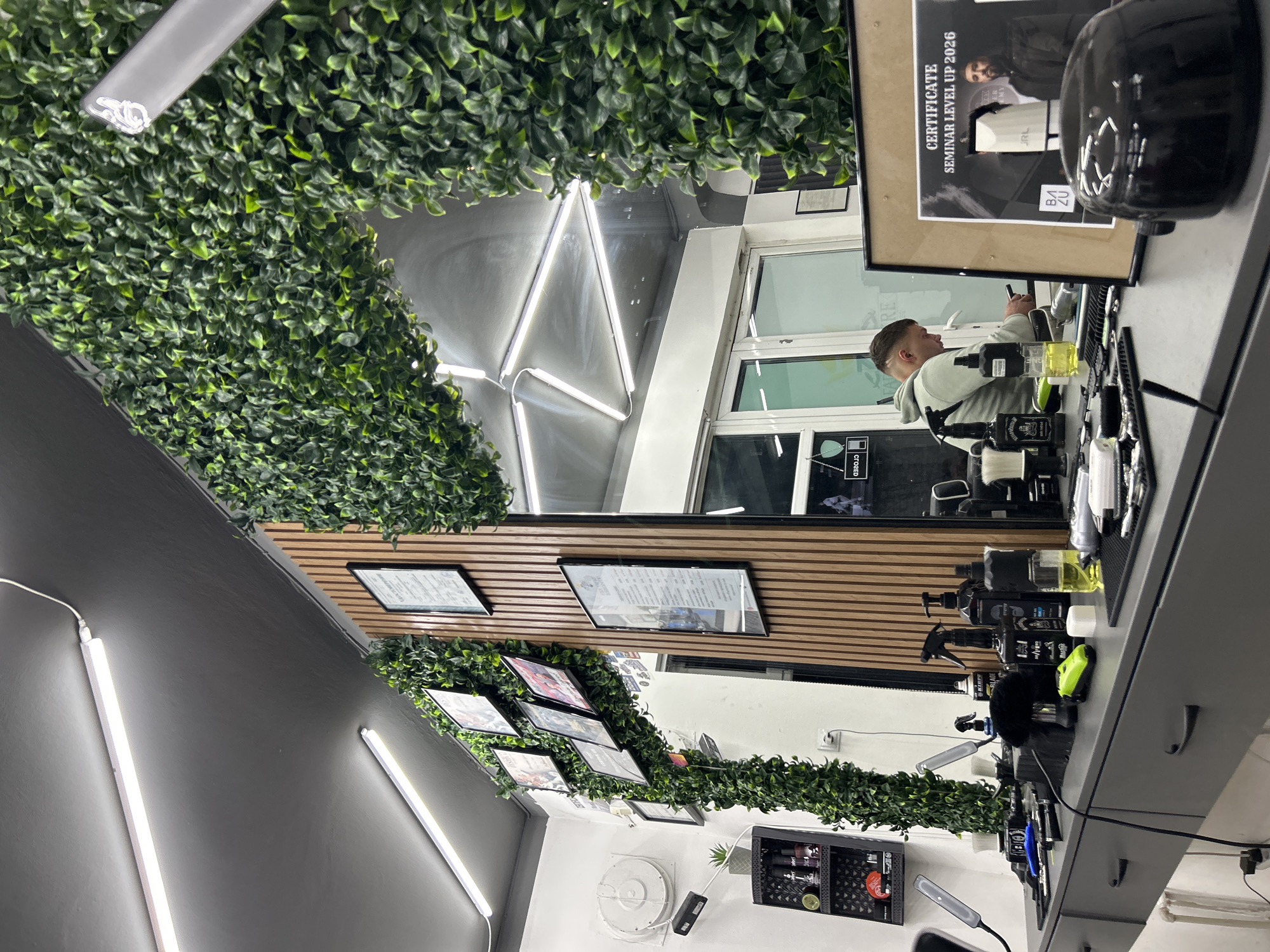 The image depicts the interior of a barbershop located in Buhovo, Bulgaria. The camera's viewpoint is from behind the barber's workstation, facing a large mirror. The mirror reflects the opposite side of the shop, showcasing a client seated in a barber chair. The client is wearing a light-colored hoodie. The walls are painted gray and adorned with linear LED lighting. Above the mirror, artificial green foliage is arranged, creating a decorative border. Below the mirror, framed certificates and framed photographs are mounted on a wood-paneled section of the wall. The workstation is cluttered with barbering tools and products, including clippers, bottles, and brushes. A framed certificate with the text "CERTIFICATE SEMINAR LEVEL UP 2026" is placed on the workstation. The overall environment appears modern and well-lit.