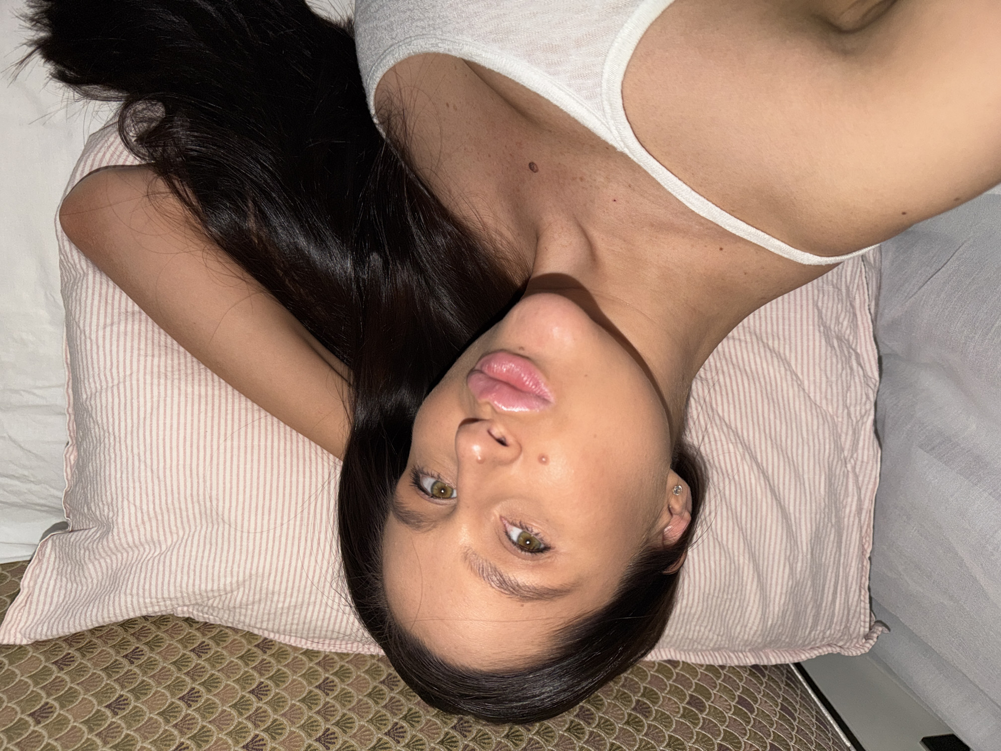 A woman with long, dark hair is lying on a striped pillow on a patterned bedspread, wearing a white tank top. The location is not identifiable as Khimki, Russia.