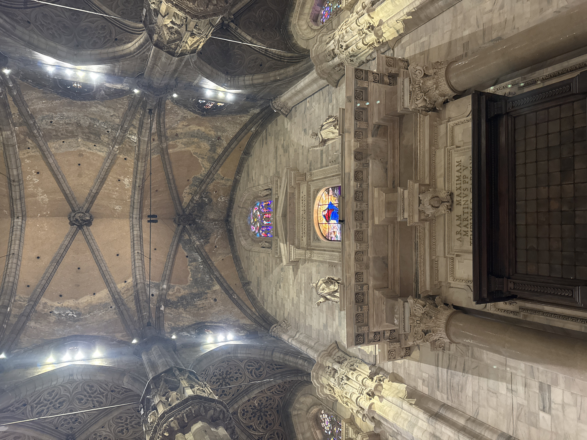 The image shows a low-angle view looking up at the ornate interior of the Milan Cathedral, also known as the Duomo di Milano, in Milan, Italy. The architecture features a high vaulted ceiling with intricate gothic ribbing and decorative stonework. Sunlight streams through stained-glass windows located high on the walls, casting colorful light. Several statues and carved reliefs are visible, as well as large marble columns. The overall impression is one of grandeur and historical significance.
