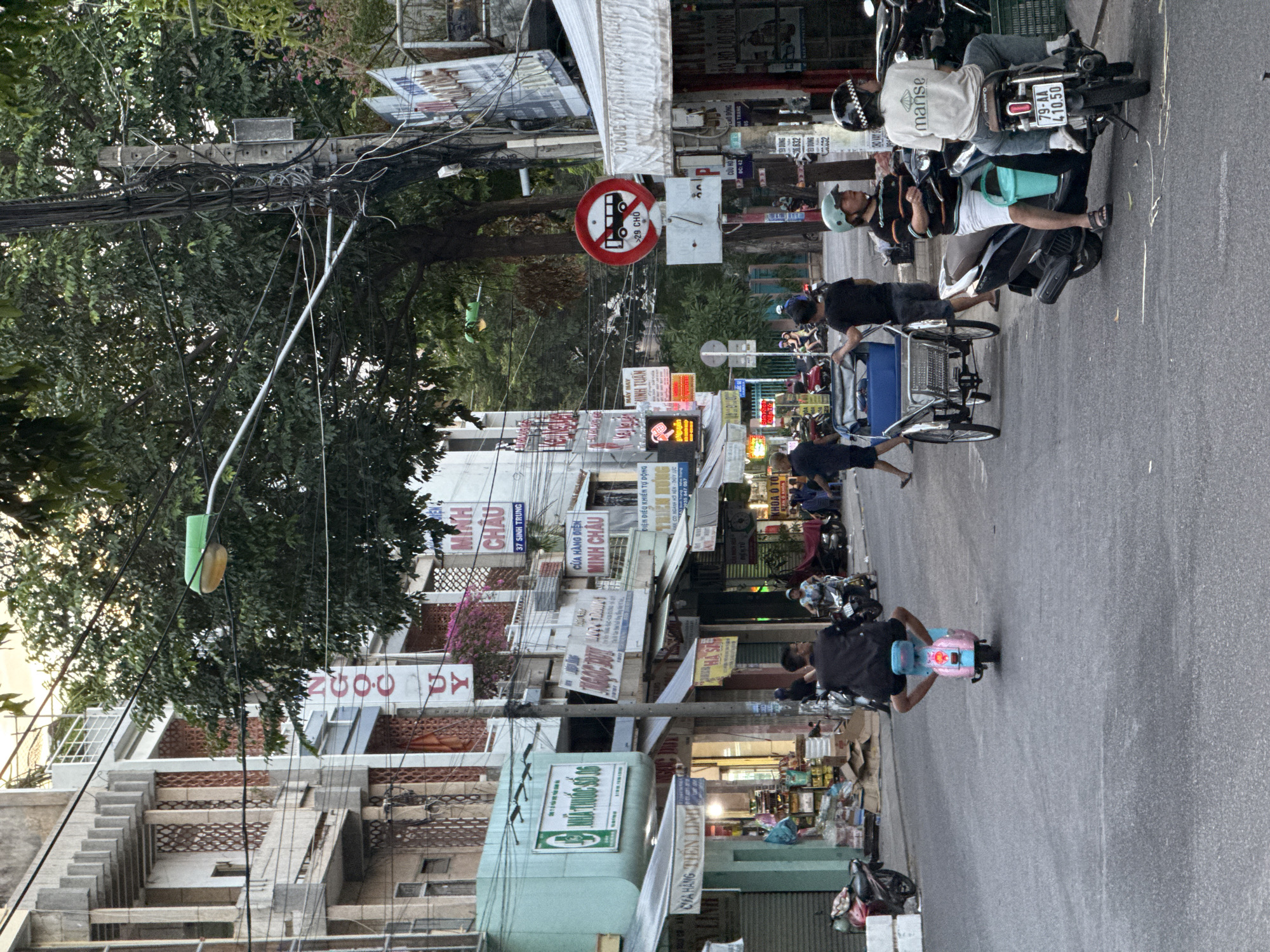 The image shows a street scene in Nha Trang, Vietnam. The street is lined with buildings of varying architectural styles, many of which appear to be commercial establishments with signs in Vietnamese. Several people are visible, mostly on motorbikes and scooters, which are a common mode of transportation in Vietnam. One person is riding a pink and blue scooter, another is on a black scooter with a passenger wearing a white t-shirt with the word "manse" on it and a checkered helmet. A three-wheeled cycle rickshaw is also present, being pushed by a person in a black t-shirt. Traffic signs are visible, including a circular red and white sign indicating a restriction on buses. Power lines are strung overhead, typical of urban environments in Vietnam. The general atmosphere suggests a bustling street during daylight hours, with the sun casting some shadows. The buildings on the left have intricate concrete facades and balconies, while those on the right have awnings and more standard shopfronts. Trees with lush green foliage are interspersed with the buildings and overhead wires. The road surface is asphalt.