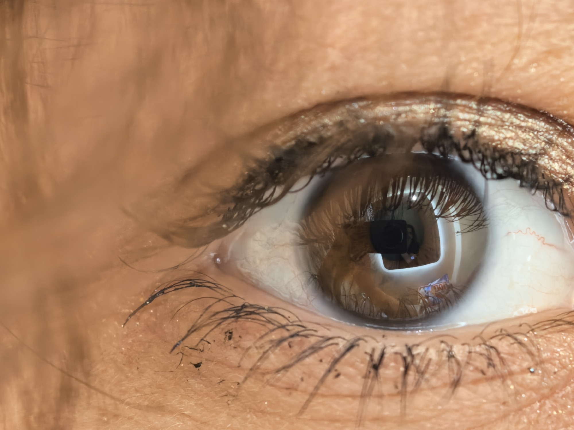 A close-up, eye-level, macro photograph captures the right eye of an individual. The eye is a deep brown, with visible eyelashes on both the upper and lower lids. Shimmering makeup is applied to the eyelids, and some loose glitter particles are visible on the skin. The sclera, or white part of the eye, shows a network of fine red blood vessels. The reflection in the pupil shows a black object, possibly a camera lens, and a blurry, abstract image of blue and orange colors. The surrounding skin appears to be of a warm, tanned complexion, with subtle textures and some fine hairs. The lighting is focused on the eye, creating a high contrast and highlighting the details. The background is a soft, out-of-focus blur of warm tones, suggesting an indoor or studio environment.