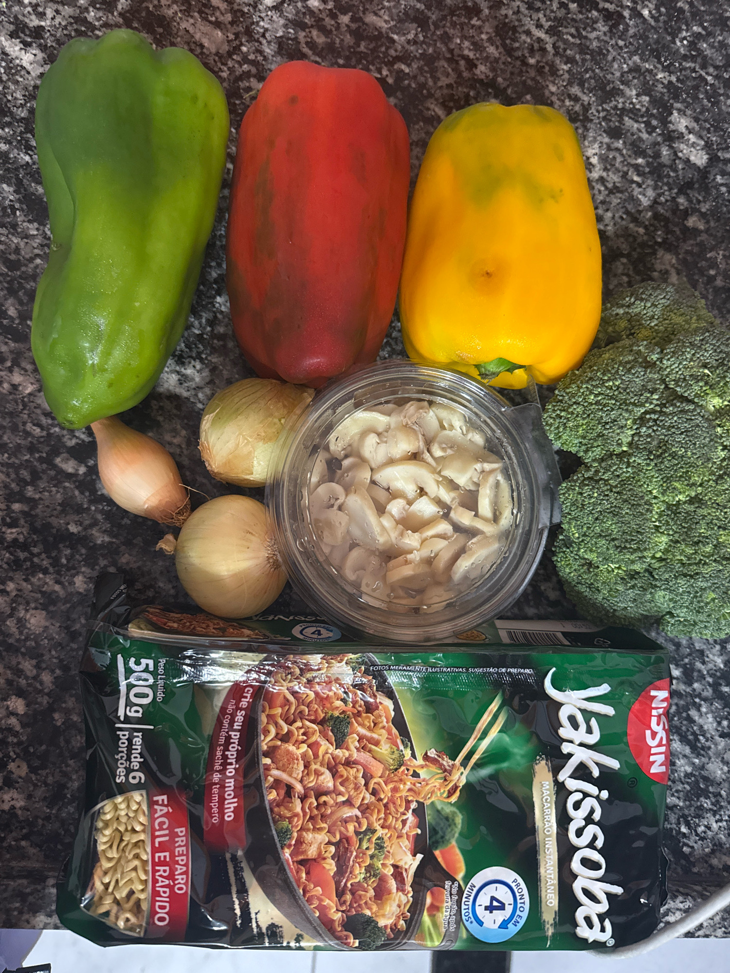 The image displays an overhead view of various ingredients arranged on a speckled gray countertop. The ingredients include a green bell pepper, a red bell pepper, a yellow bell pepper, a head of broccoli, two small onions, a shallot, a clear plastic container filled with sliced mushrooms, and a package of Nissin Yakisoba instant noodles. The Yakisoba package is primarily green with white text and an image of the prepared dish. The ingredients are neatly organized, suggesting preparation for a meal. The lighting is even, and there are no discernible people or actions. The location is not identifiable as being in the city of Nilópolis, Brazil, as there are no distinctive urban features present.