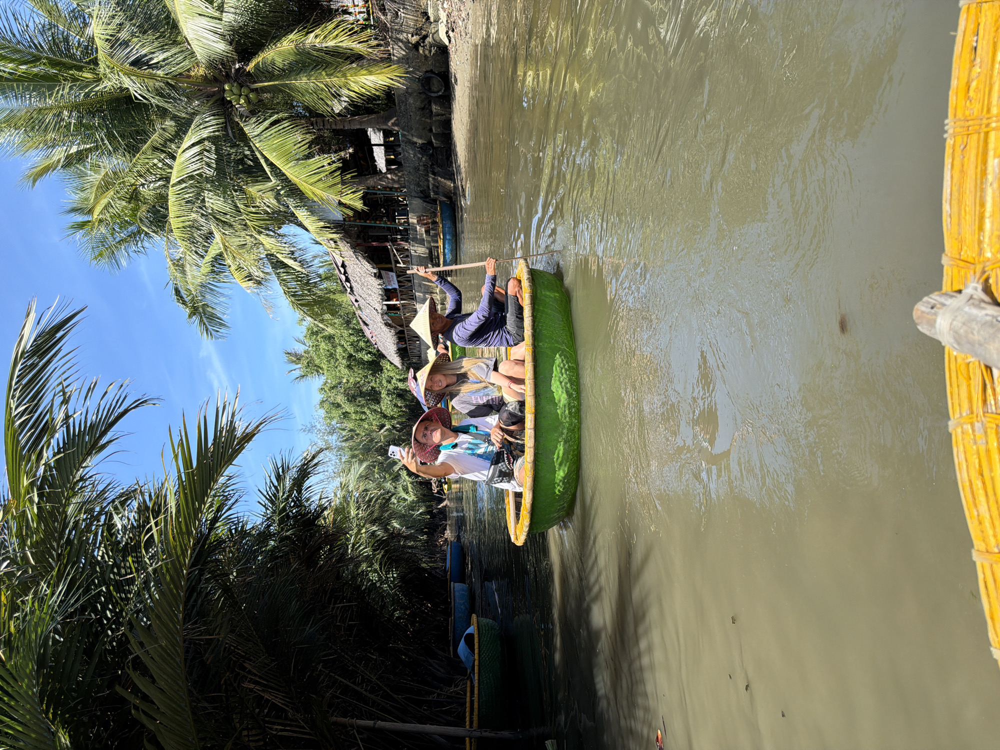 Three individuals are in a round basket boat on a river under a clear blue sky, surrounded by lush green palm trees. The person in the front-left is taking a selfie with a smartphone while wearing a conical hat and a white t-shirt. Beside them, a person with long blonde hair is also wearing a conical hat and a grey t-shirt. The third person, at the back of the boat, is wearing a dark long-sleeved shirt, dark shorts, and a conical hat, actively rowing the boat with a wooden oar. The boat has a green exterior and a yellow interior. In the background, there are more basket boats, thatched-roof structures, and a sandy bank with vegetation. The water is murky brown. The scene appears to be in the area of the Hoi An river in the city of Đà Nẵng, Vietnam.