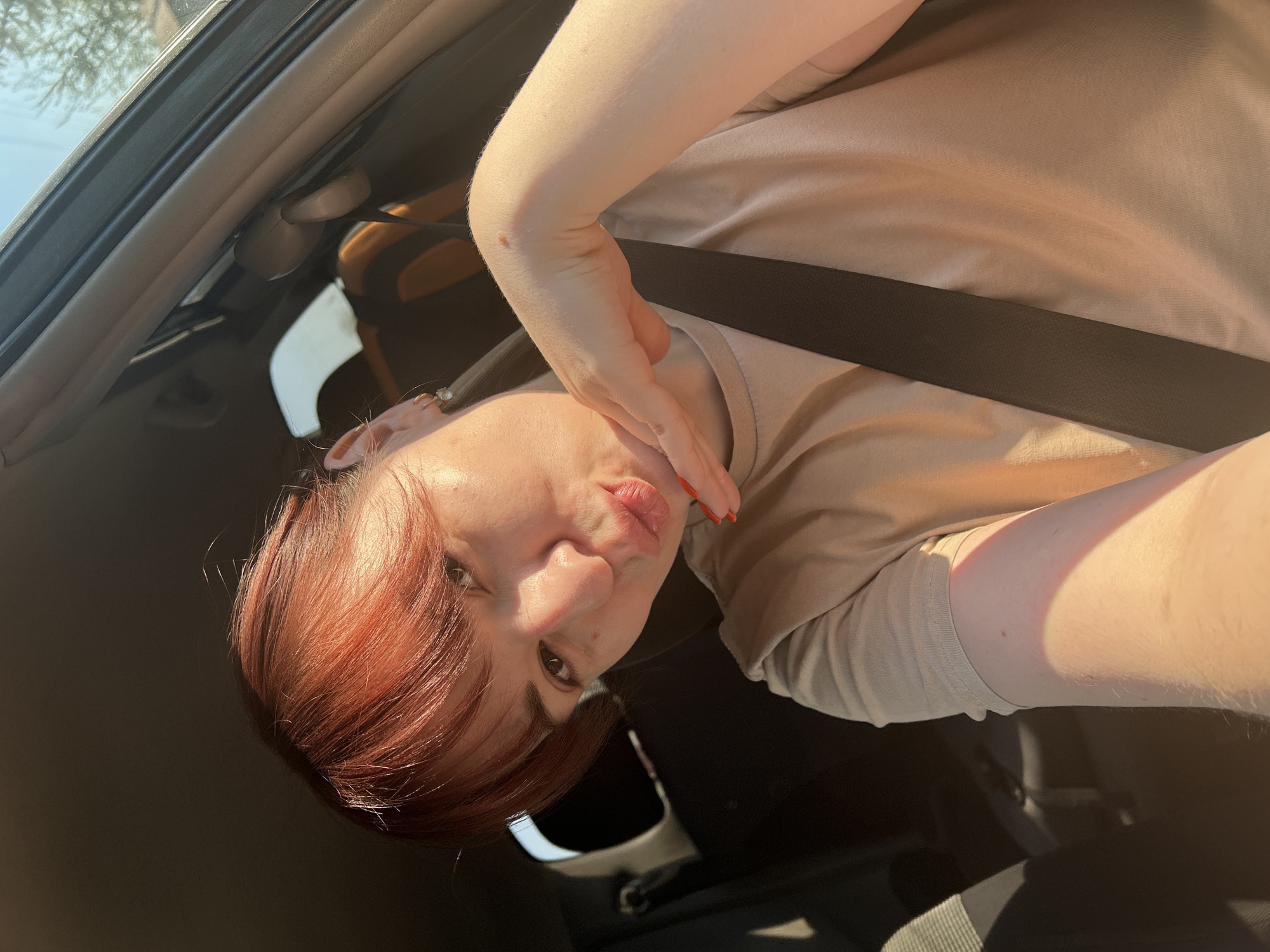 A person with red hair is sitting in a car, wearing a tan shirt and a seatbelt. They are holding their hand near their face and looking at the camera. The inside of the car is visible, including the dashboard and the roof. The location is not discernable from the image.