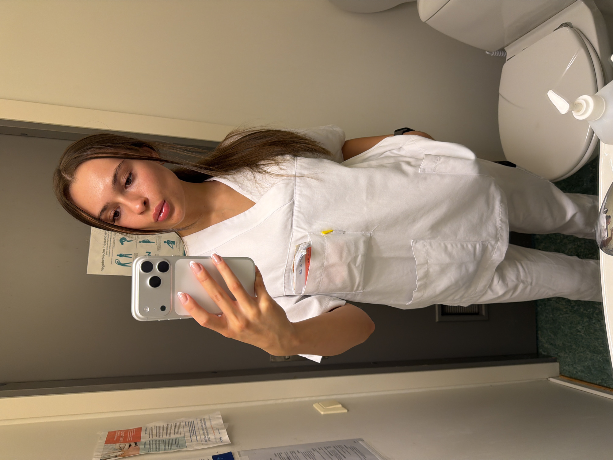 A young woman with long brown hair pulled back in a ponytail, wearing white medical scrubs, takes a selfie in a bathroom. She is holding a silver iPhone in her left hand, the screen reflecting a portion of the room. Her right hand is in her pocket. The bathroom appears to be in a medical facility, indicated by the scrubs and a poster on the wall displaying exercise diagrams. To her right, a white toilet with the lid down is visible, along with a bottle of hand soap on its tank. The floor has a green, speckled pattern. The image is captured from a slightly low angle, looking up at the woman and her reflection. The specific location in Harstad, Norway, is not discernible from the visual information provided.