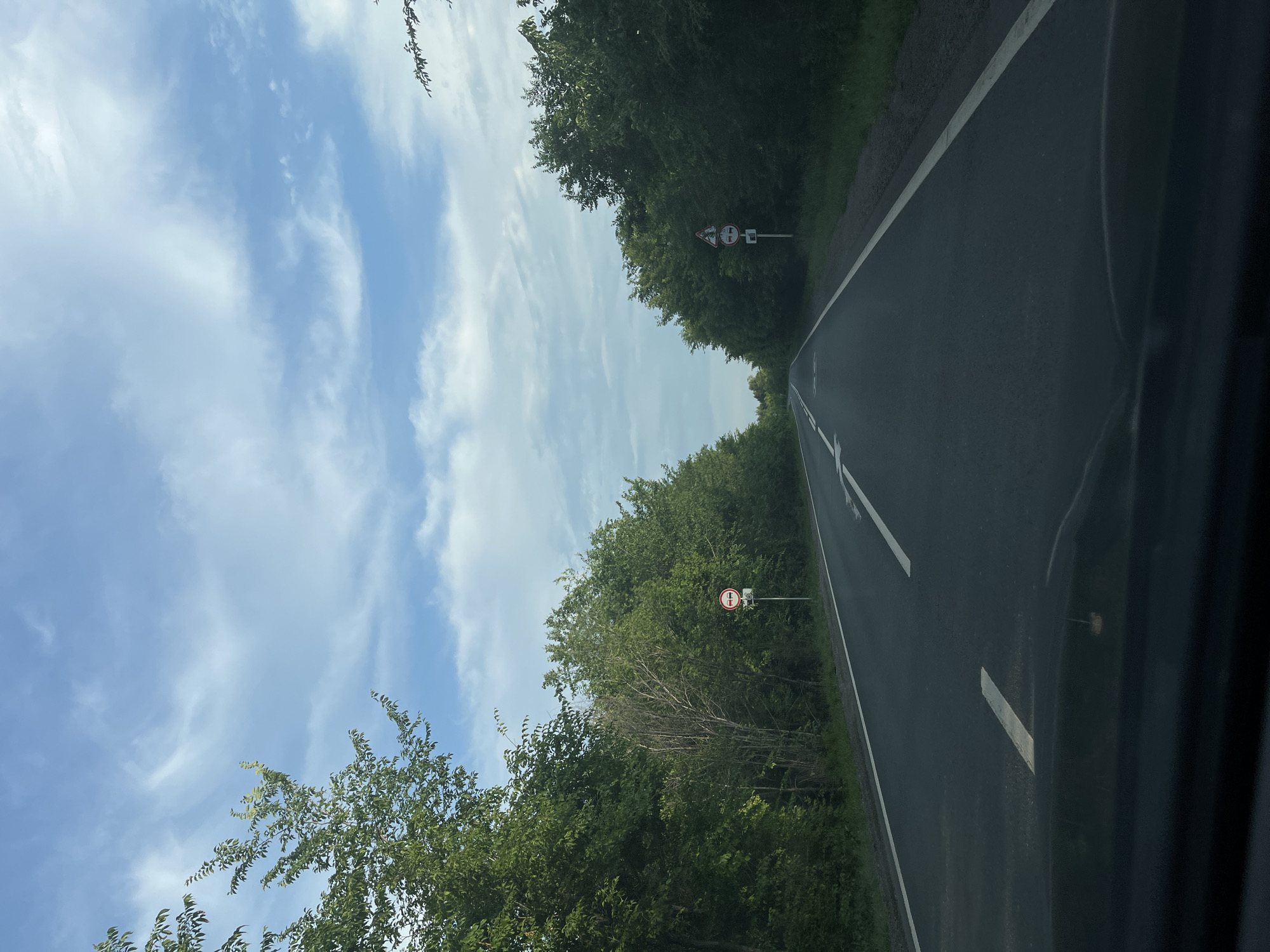The image depicts a two-lane asphalt road bordered by dense green trees on both sides. The sky above is blue with scattered white clouds. Two road signs are visible on the right side of the road, and one on the left. The sign on the left is round with a red border and a horizontal bar across it, indicating a prohibition. The signs on the right appear to be a triangular warning sign followed by a round sign with a red border and a horizontal bar, also indicating a prohibition. The perspective suggests the photo was taken from inside a vehicle driving on the road. There are no people or animals visible in the scene. The environment appears to be rural or a highway with heavy tree cover.