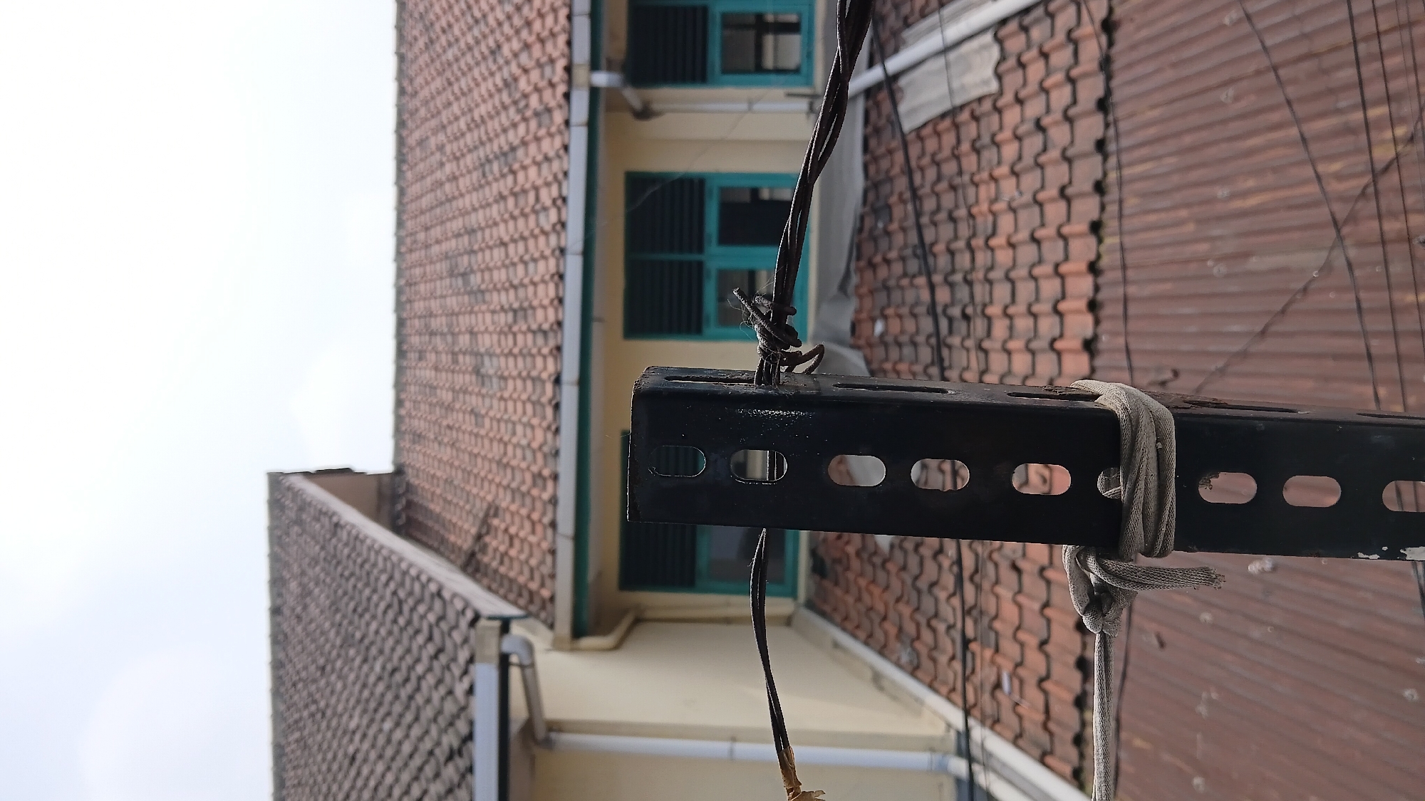 The image shows a close-up of a black metal post in an outdoor urban setting, likely a rooftop or elevated area. The post has several rectangular cutouts and is weathered with some peeling paint. Two wires are attached to the post; one is a thick, dark wire secured with a knot to the left side of the post. A thinner, lighter-colored rope is also tied around the post in the lower section. In the background, there are tiled roofs of buildings, some yellow walls, and windows with teal-colored shutters. The sky is overcast. The scene depicts a utilitarian structure against a backdrop of residential or commercial buildings. The context of Bandung City, Indonesia, suggests a tropical or subtropical urban environment.