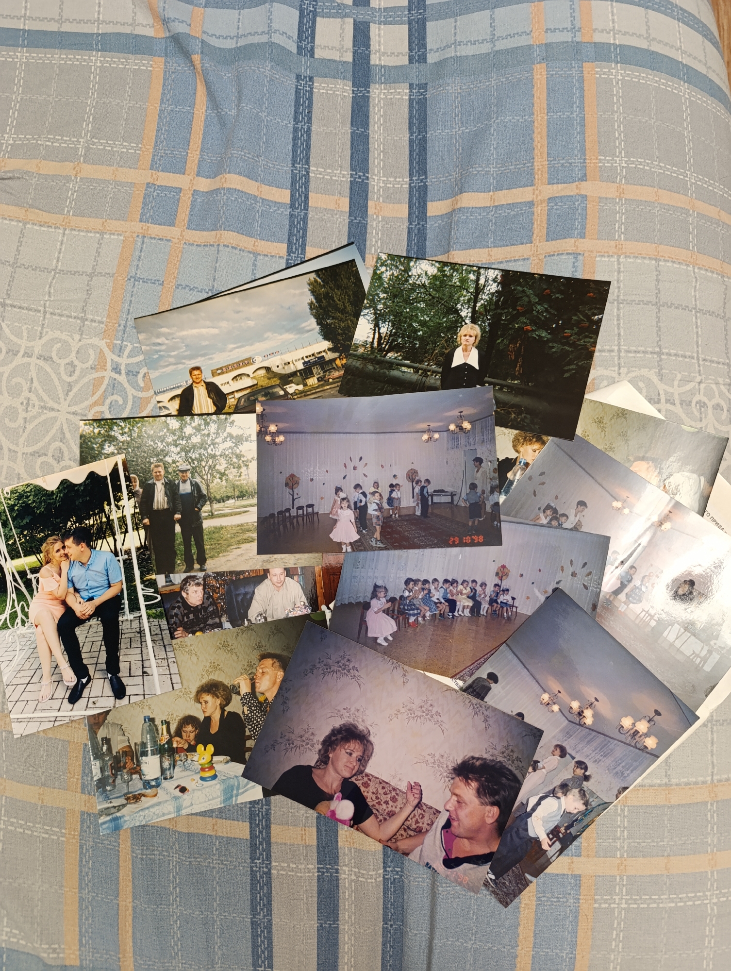 The image shows a collection of photographs scattered on a patterned bedspread. The photographs depict various scenes, including people posing outdoors, groups of children in what appears to be a classroom or daycare setting, and indoor gatherings of adults and children. The photographs show different people interacting and socializing. The environment appears to be domestic or public. There is a photograph with "29 10 98" stamped on it. The location in the city of Unknown, Unknown.