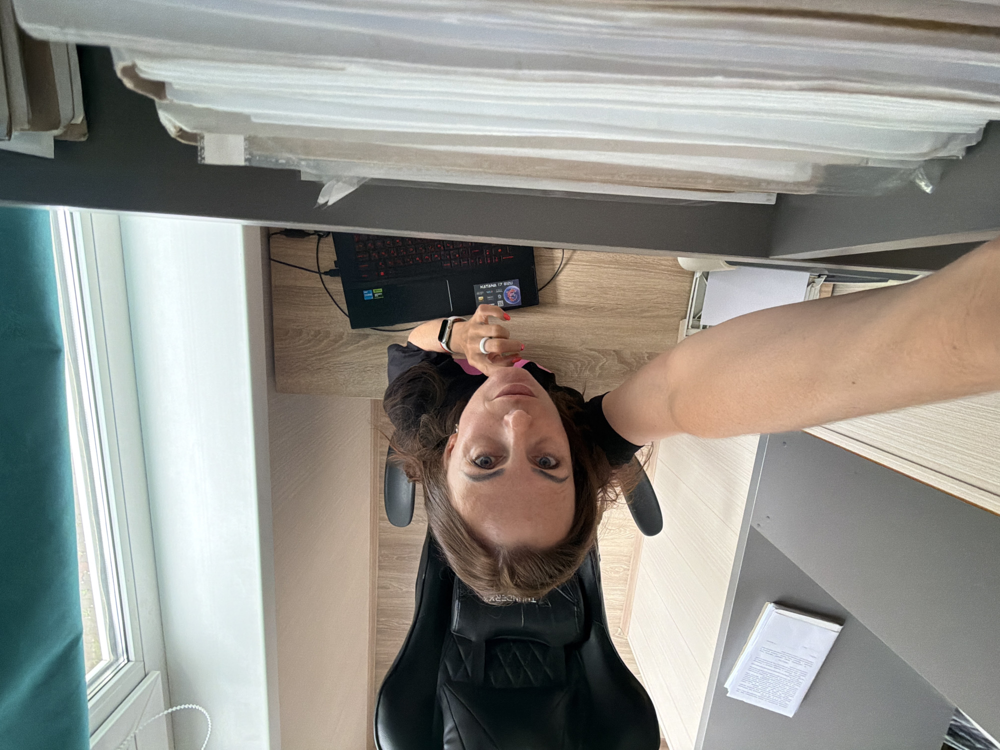 A high-angle, top-down selfie captures a woman seated in a black gaming chair, looking directly up at the camera. She has brown hair, light eyes, and wears a dark top, with pink nail polish visible on her fingers. A white ring is on her right ring finger, and a white smartwatch is on her left wrist. Her expression is neutral and attentive.
The setting appears to be an indoor office or home office. Behind her on a light wooden desk, a black laptop with a red backlit keyboard is open and plugged in. Visible text on the laptop bezel includes "KATANA 17 B13U," along with "Intel Core i5" and "GeForce RTX" logos.
Above her head, a shelf is laden with numerous stacks of white papers or documents, stored horizontally. To her left, a window with white frames and a teal curtain provides natural light. On a lower shelf or surface to her right, another stack of documents with visible text is present. The gaming chair features the brand "THUNDERX3" on its headrest. The flooring is a light-colored wood. The overall impression is a personal workspace.