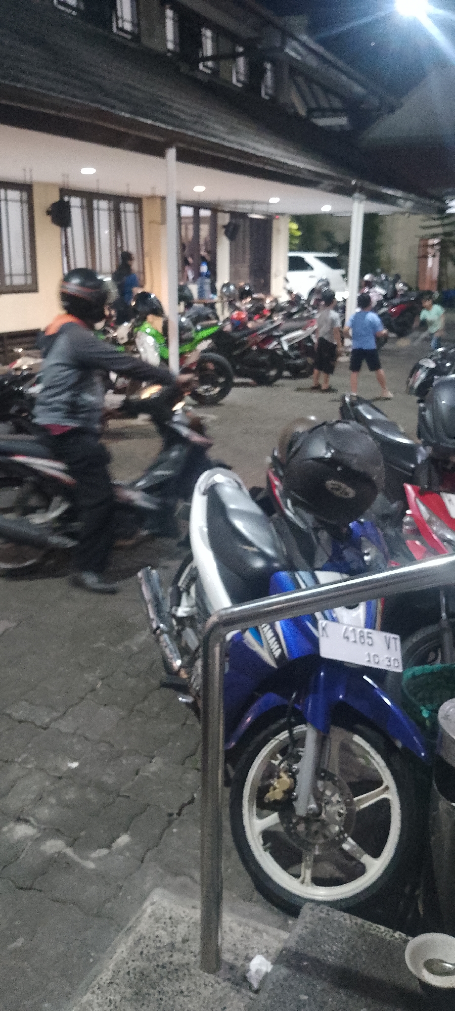 The image shows an outdoor area, likely a parking lot, at night. In the foreground, a blue and white Yamaha motorcycle is prominently displayed, with its license plate reading "K 4185 VT 10 30". Several other motorcycles are parked in the background, with some people visible among them. The setting appears to be in front of a building with a covered walkway or overhang. The ground is paved with interlocking concrete blocks. The overall atmosphere suggests a casual gathering or a stop at a roadside establishment in Kudus, Indonesia.