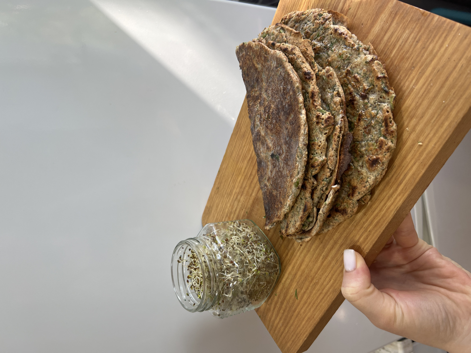 A light-skinned hand, with short nails painted in a nude-pink polish, grips the right edge of a rectangular wooden cutting board in the lower-left quadrant of the frame. On the light-colored wooden board, a stack of approximately five to six round or oval-shaped, savory flatbreads or crepes, mottled brown and green from herbs or vegetables, is arranged in the middle-right. The flatbreads show various degrees of browning and slight charring from cooking. To the upper-left of the flatbreads, positioned between them and the holding hand, sits a clear, squat glass jar, possibly hexagonal, filled with a dense mass of light green and white sprouted seeds, some still attached to their dark husks. The background is a plain, light off-white or very light gray wall. A distinct, diagonal beam of sunlight streams from the upper right, casting a shadow across the upper-right portion of the wall. The scene is indoors in the city of Unknown, Unknown.