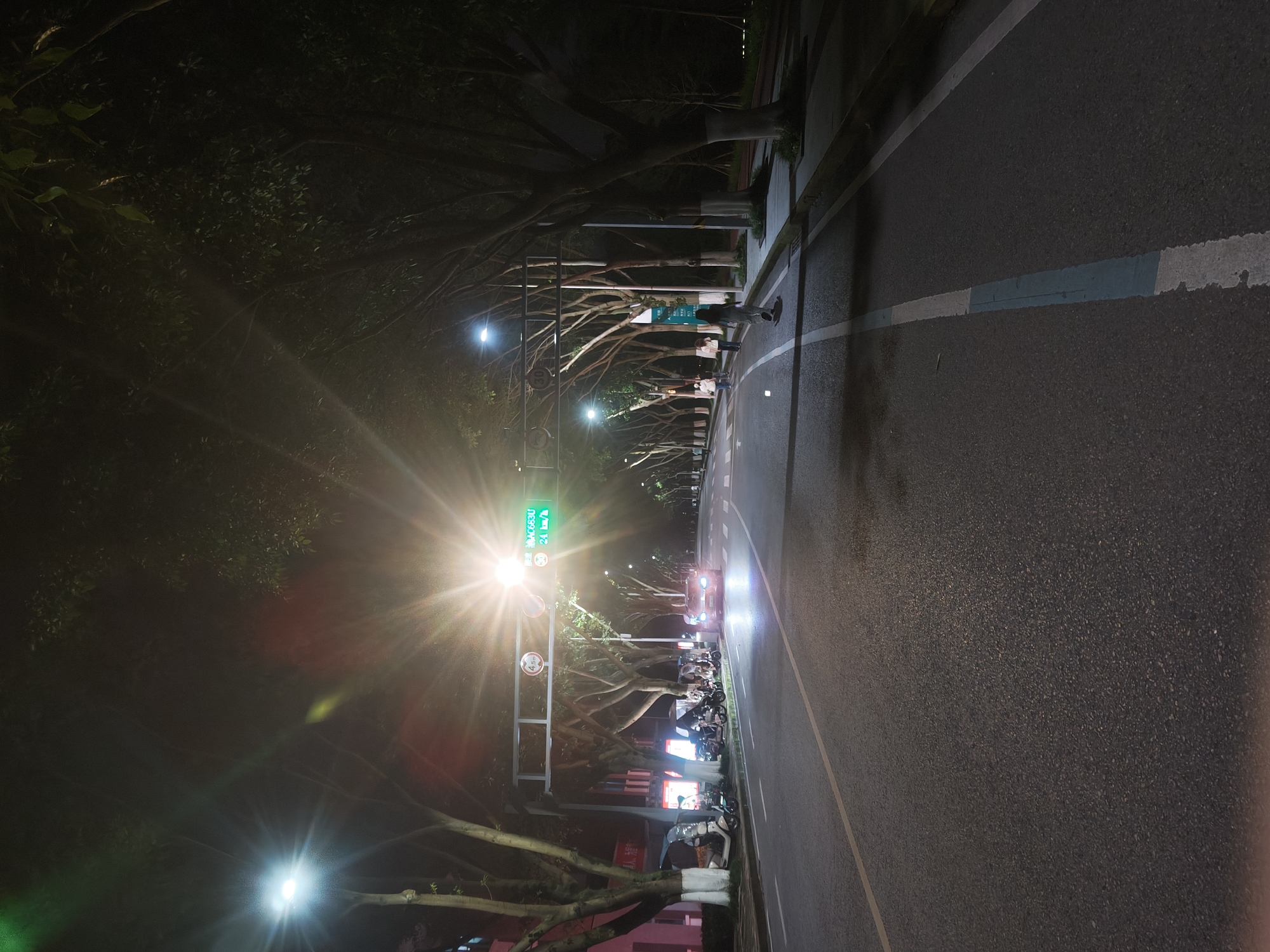 A nighttime, eye-level, wide shot captures a street scene in the Chongqing High-tech Zone, China. The road, paved with asphalt, is illuminated by streetlights and the headlights of a car. Trees with white-painted lower trunks line both sides of the road, their branches forming a canopy overhead. In the foreground, a white line demarcates the edge of the road. A parked line of electric scooters is visible on the left side of the street. Further down the road, a car with its headlights on is approaching. Several people are walking on the sidewalk on the right side of the street. A traffic light with a speed limit display of 24 km/h hangs over the road. The overall impression is a well-lit urban street at night.
