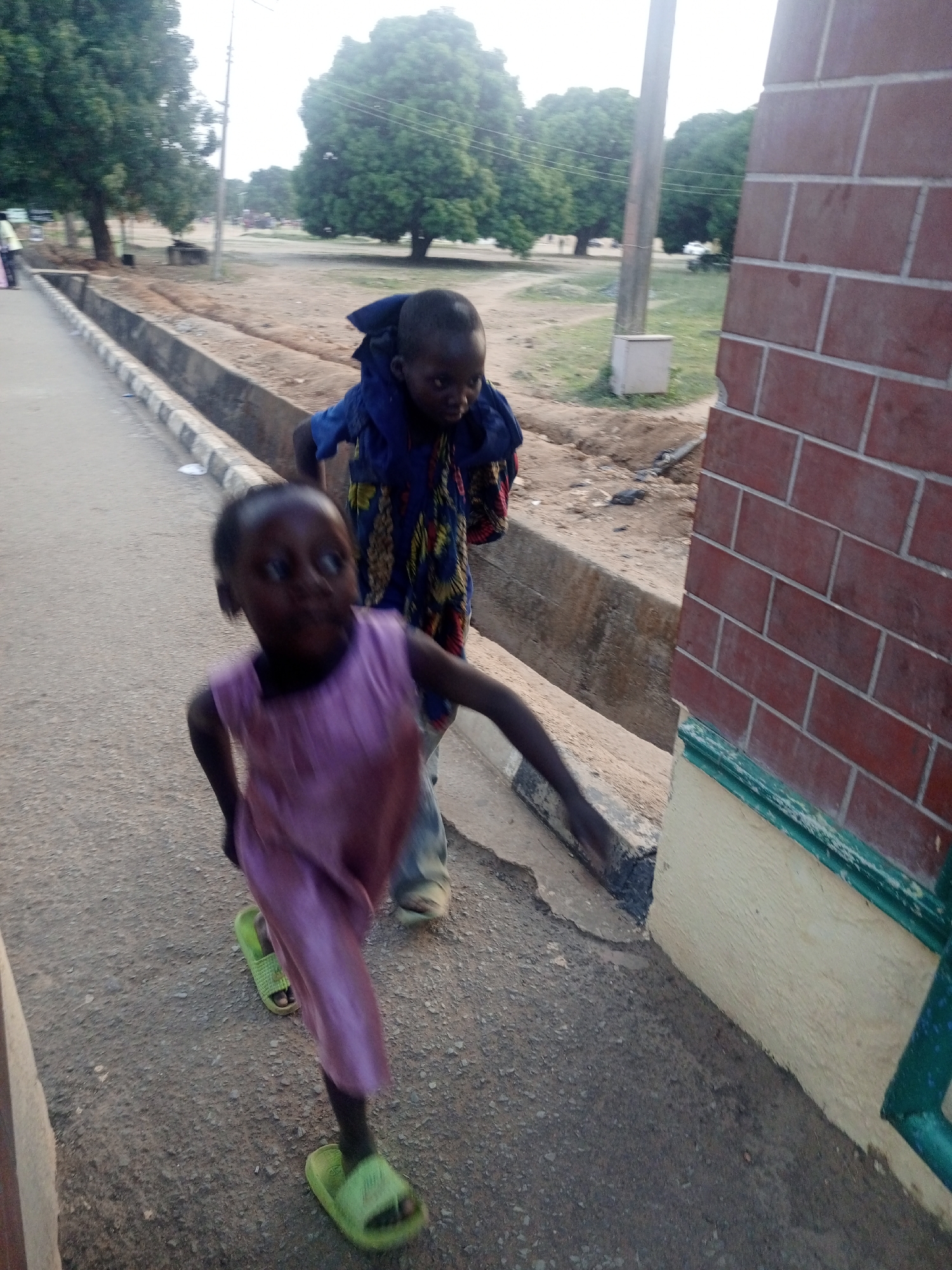 The image shows two young children walking or running along a paved path in Keffi, Nigeria, captured likely in the late afternoon or early evening. In the foreground, a child with dark skin, whose face is blurred by motion, wears a light purple sleeveless dress and bright green slip-on sandals. Their arms are bent, indicating movement.
Behind this child and slightly to the right, a second child with dark skin is visible, wearing a blue garment with distinct yellow and red patterns, possibly a wrap or patterned top over a blue shirt. This child appears to be carrying something on their back, covered by the blue fabric, and has a neutral expression.
The children are on a path made of asphalt or concrete, which has some small debris. To their left, a concrete curb with black and white stripes separates the path from a concrete drainage ditch. On the right side of the frame, a red brick wall with white mortar rises vertically, featuring a light yellow base and a dark green trim or railing section.
In the background, several large, green trees are prominent, suggesting a natural or semi-rural environment. A utility pole stands further back, and a small, light-colored box, possibly an electrical utility box, is visible near it. The overall setting is an outdoor scene, likely a street or pathway next to a building or wall. No visible text is present in the image.